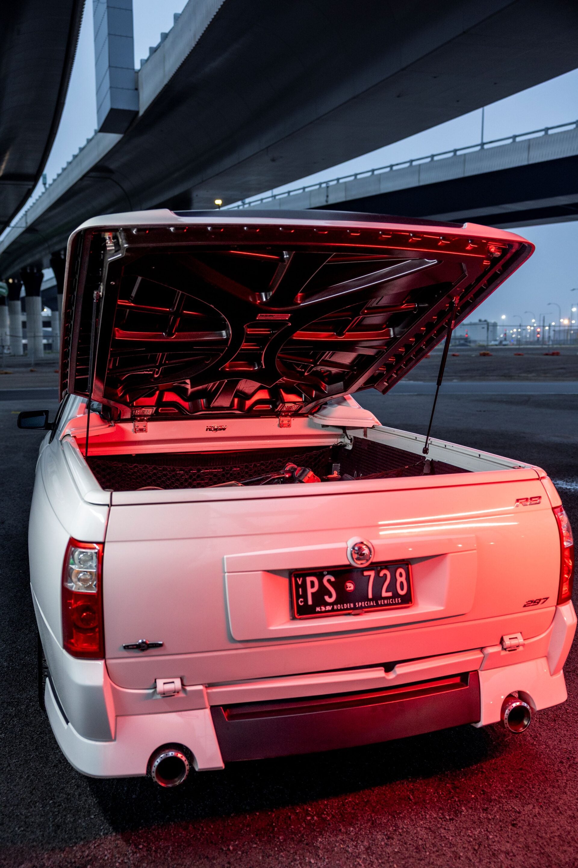 White pickup truck with the tailgate raised and red underglow lighting under concrete overpasses.