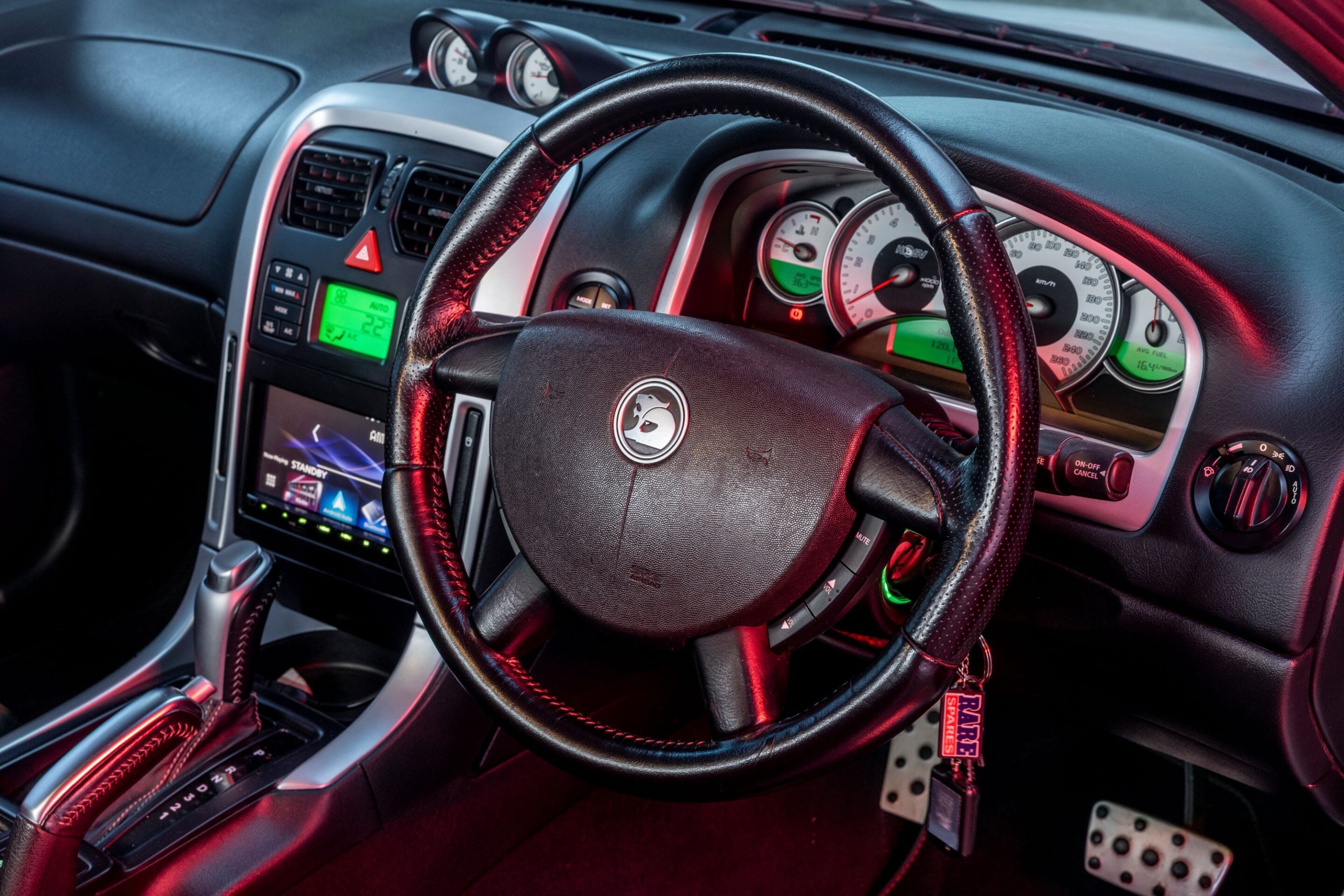 Close-up of a car's black leather steering wheel and dashboard with gauges and a green-lit infotainment display in the center console.