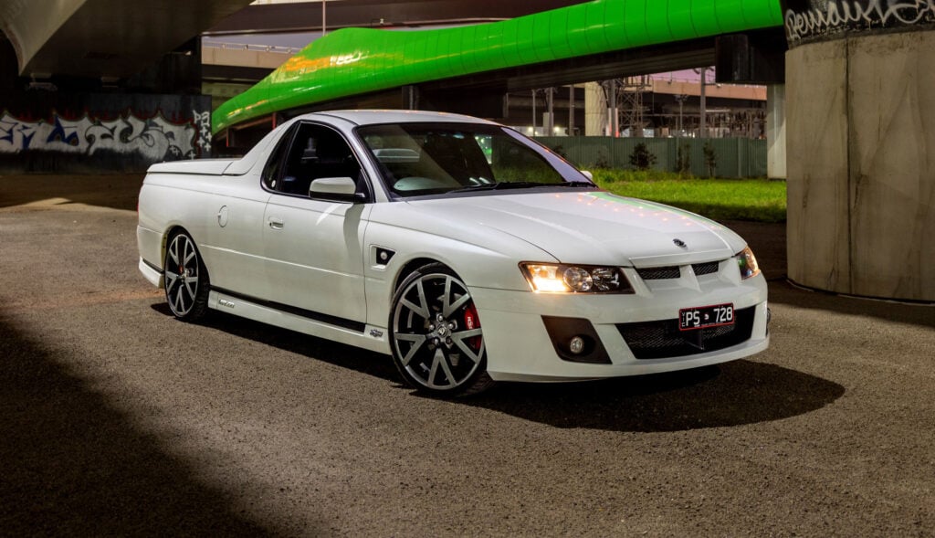 White modified pickup under a green overpass at night, front angled toward camera, custom wheels and red brake calipers visible in low light.