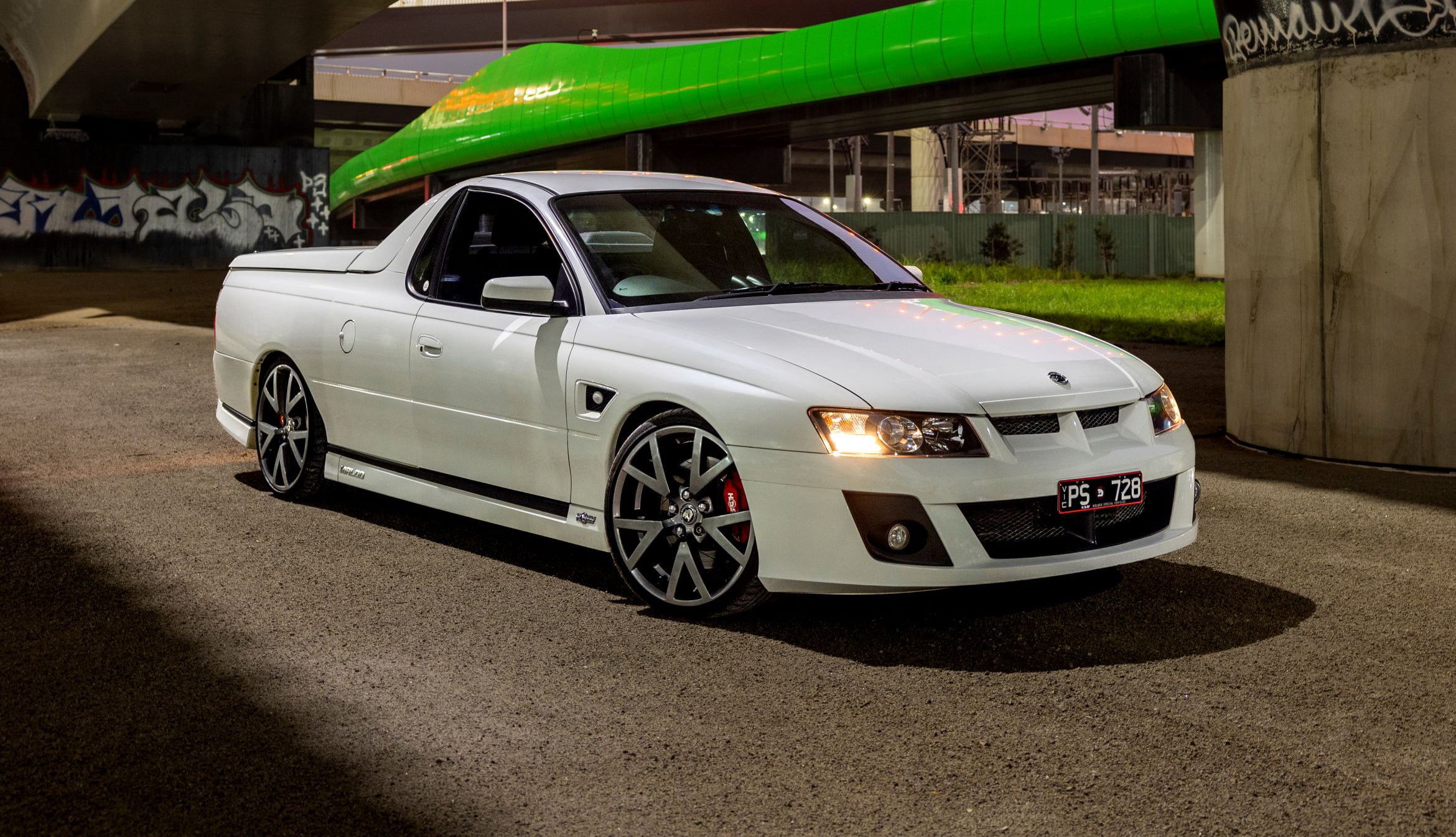 White modified pickup under a green overpass at night, front angled toward camera, custom wheels and red brake calipers visible in low light.