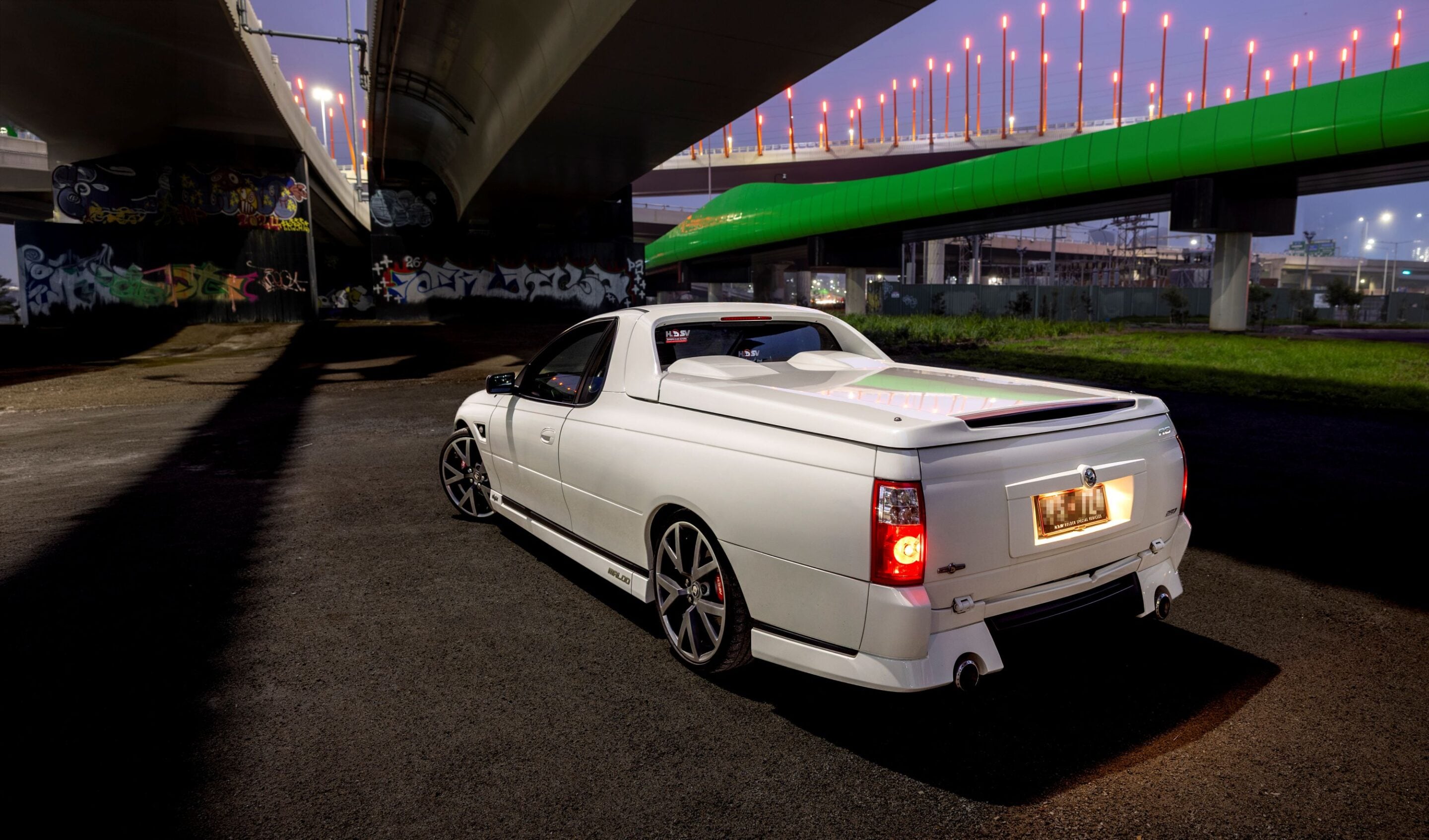 White pickup truck parked under a graffiti-covered overpass at dusk with a bright green elevated walkway above.