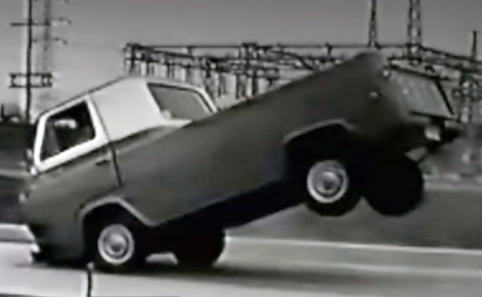 Black-and-white photo of a vintage pickup doing a wheelie, with its front wheels raised on a street.