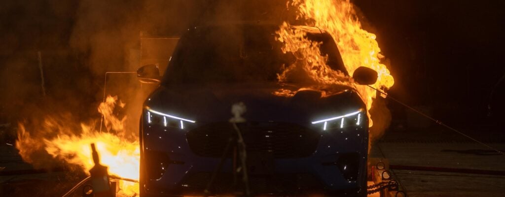 Blue car engulfed in flames at night, flames billowing from the hood and windows while the vehicle burns.