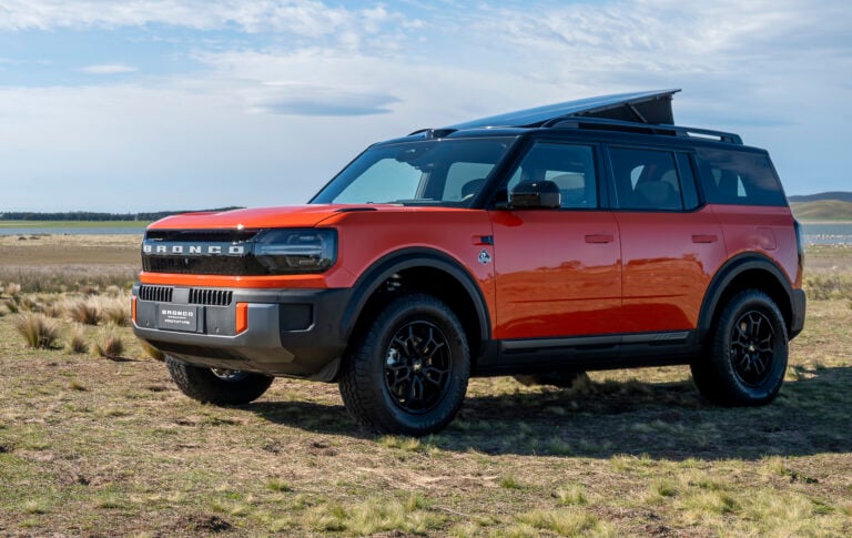 Red Ford Bronco SUV with a raised rooftop tent parked on a grassy plain under a blue sky.