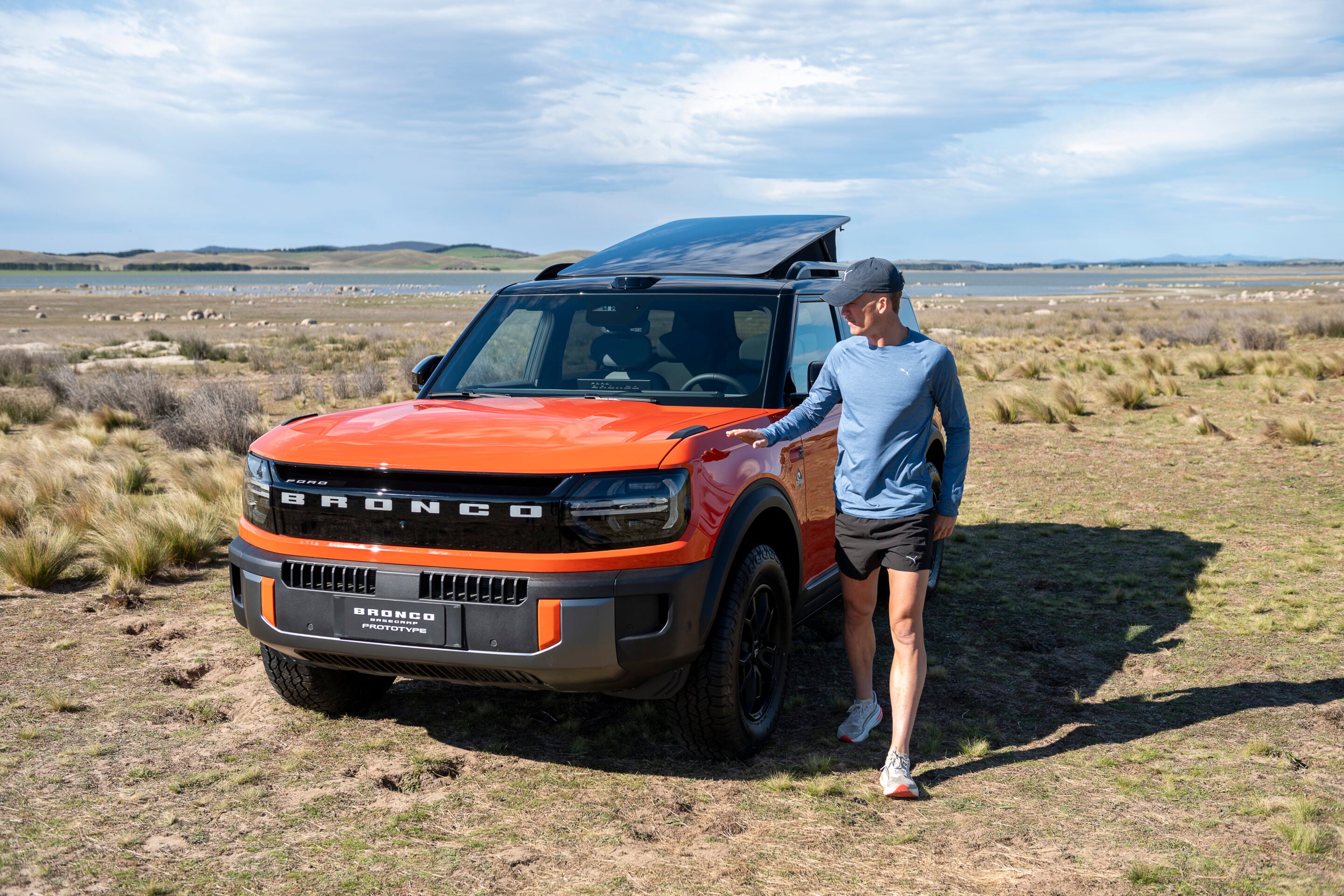 Man wearing a blue long-sleeve shirt and cap stands beside an orange Ford Bronco with its roof panel lifted, in a dry grassy landscape near water under a cloudy sky.