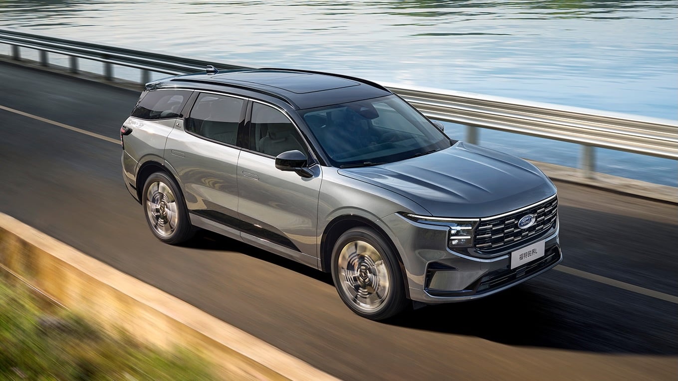 Three-quarter front view of a silver Ford SUV driving along a coastal road by a guardrail and calm water.