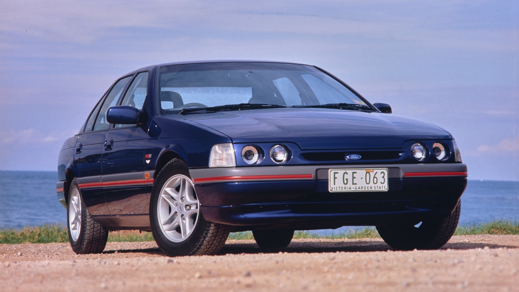 Front three-quarter view of a blue Ford sedan parked on a dirt road by the sea, license plate FGE-063.