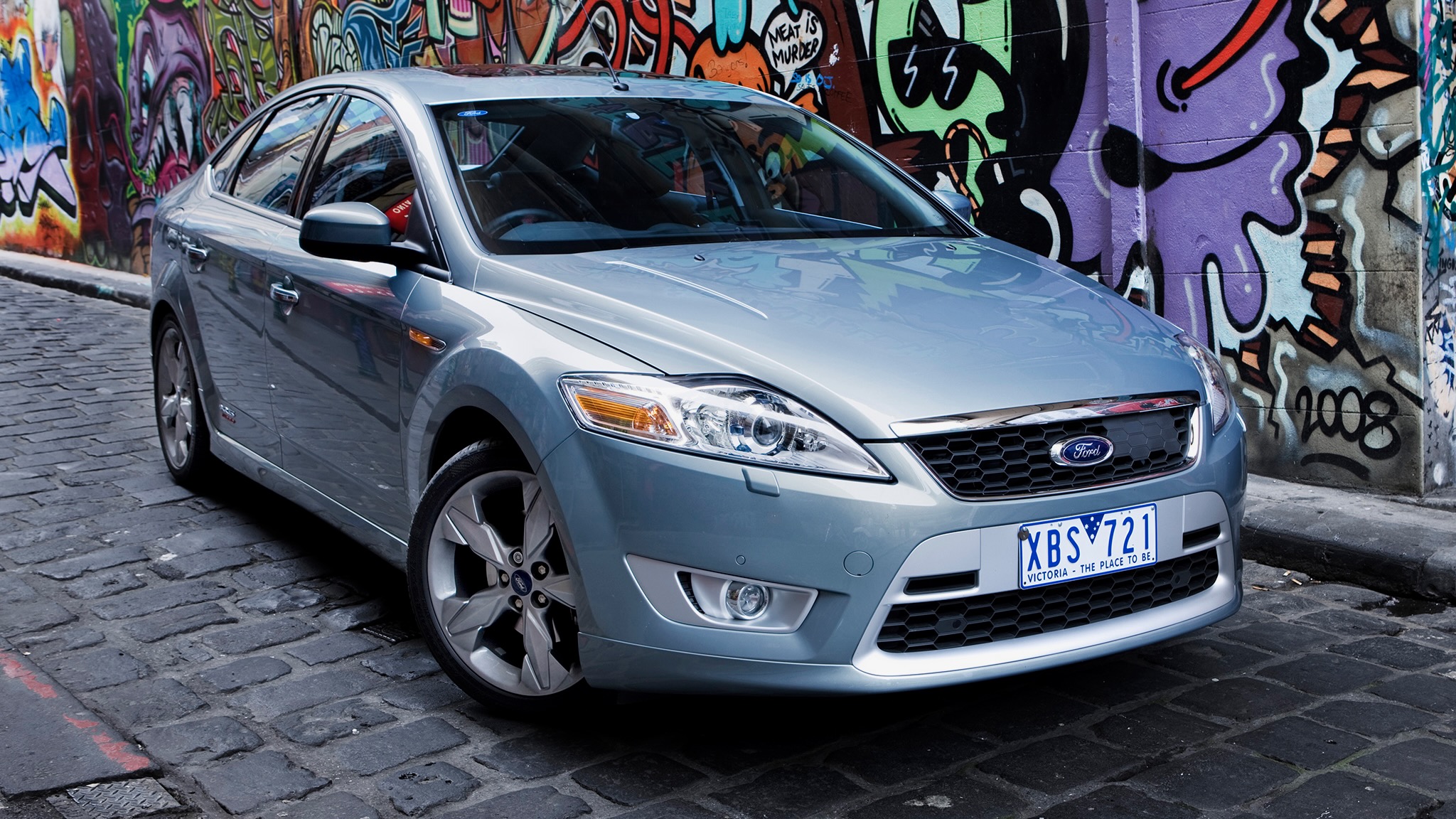 Three-quarter front view of a silver Ford sedan parked on a cobblestone street beside a colorful graffiti wall.