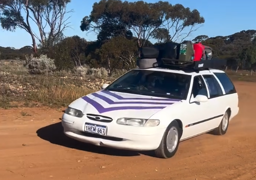 White station wagon with purple stripes on a dusty dirt road, roof rack loaded with suitcases, Australian outback in the background.