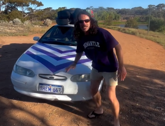Man with long hair and sunglasses beside a white car with purple chevron stripes on a dirt road near a lake.
