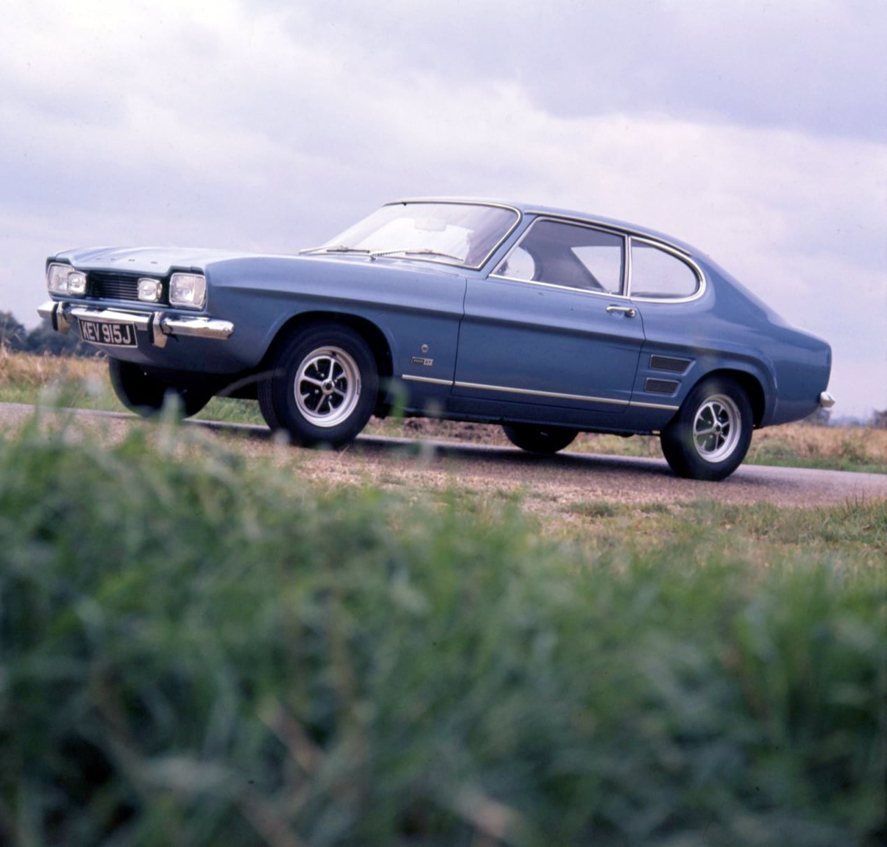 Blue vintage two‑door car on a rural road, front three‑quarter view with chrome bumper and round headlights.