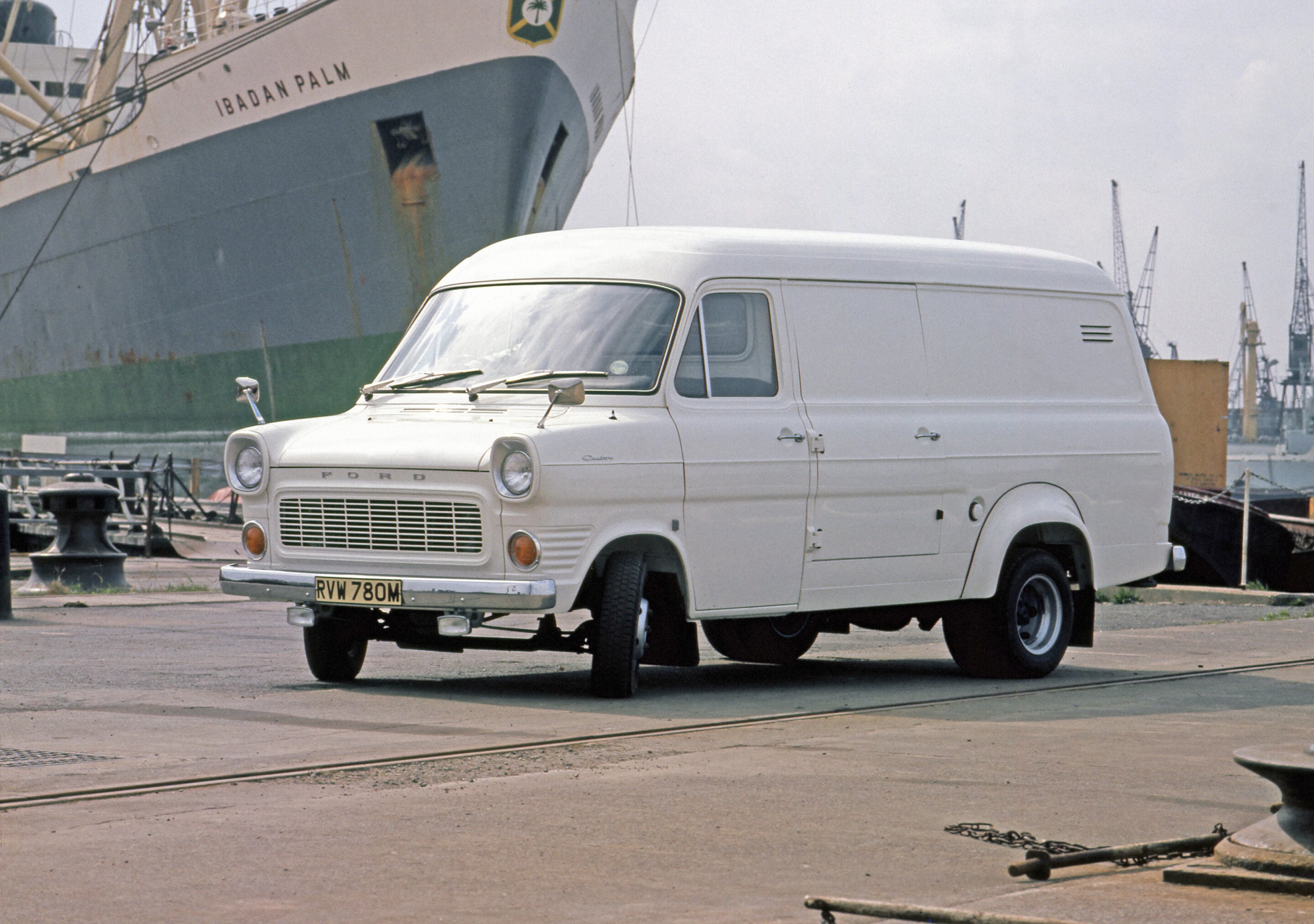 White vintage Ford van parked at a harbor, with a large ship in the background.