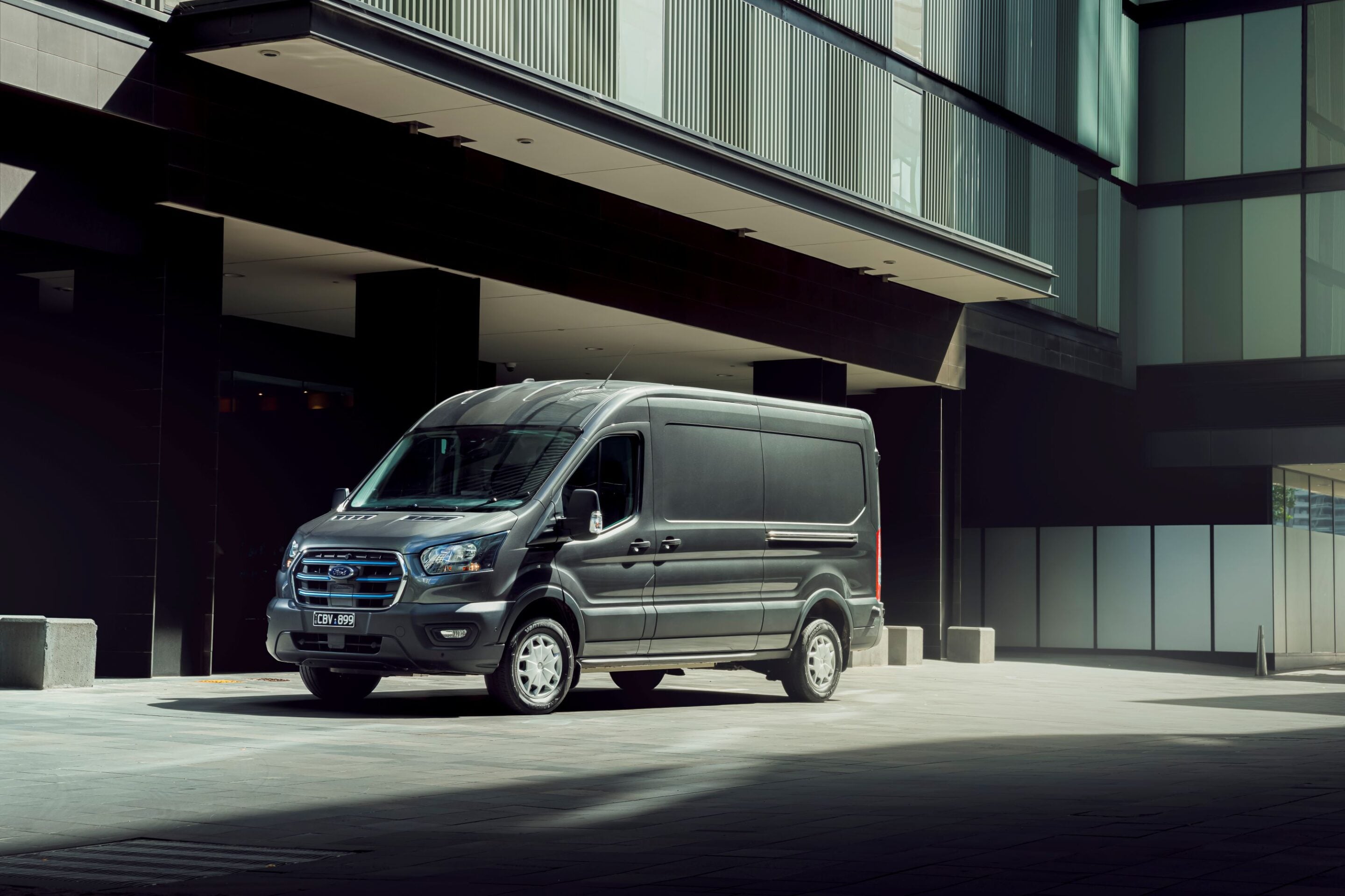 Grey delivery van parked under a modern glass-and-steel building, with large windows and tall shadows on the pavement.