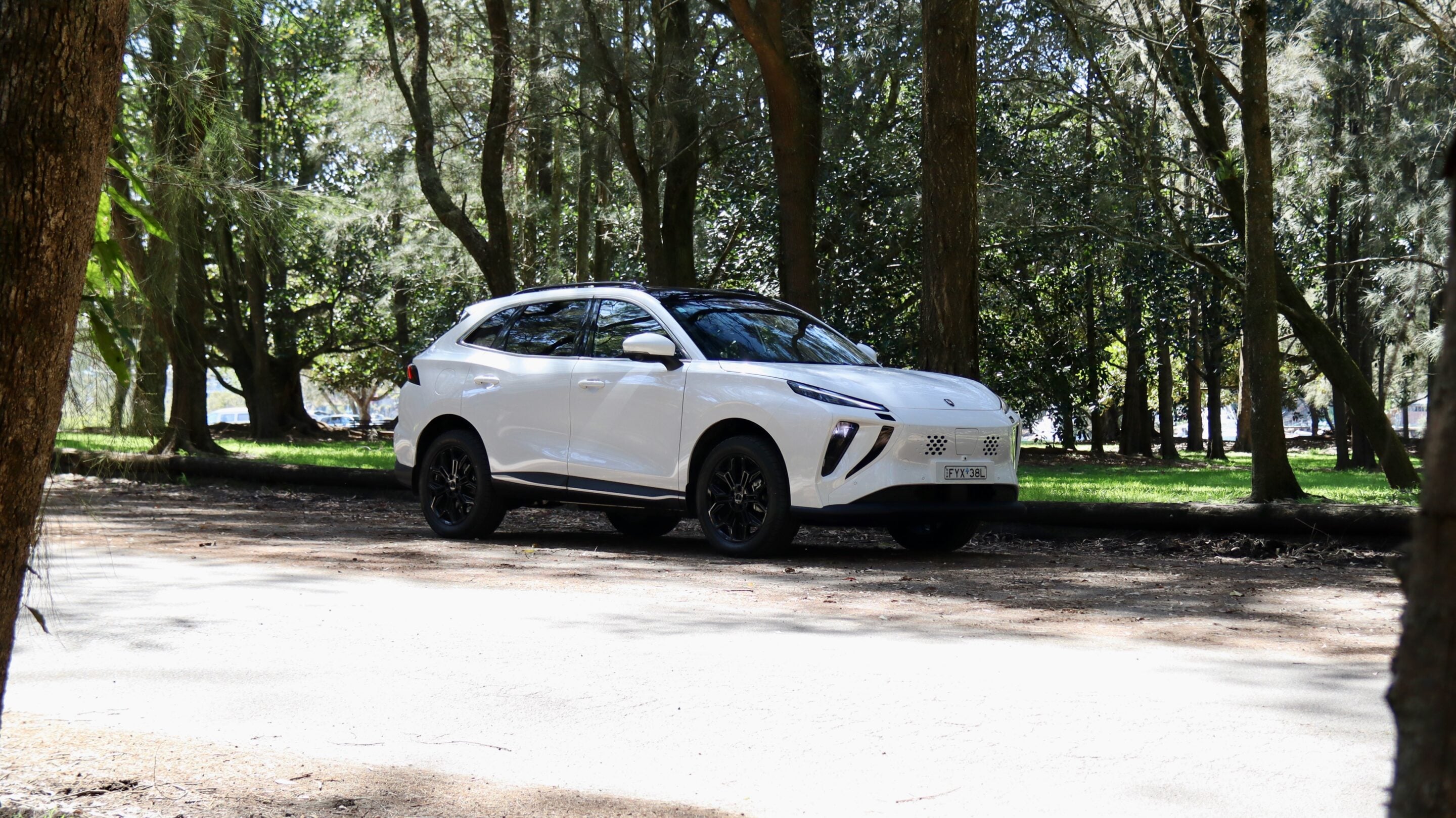 White SUV parked on a dirt path under tall trees in a shaded park.