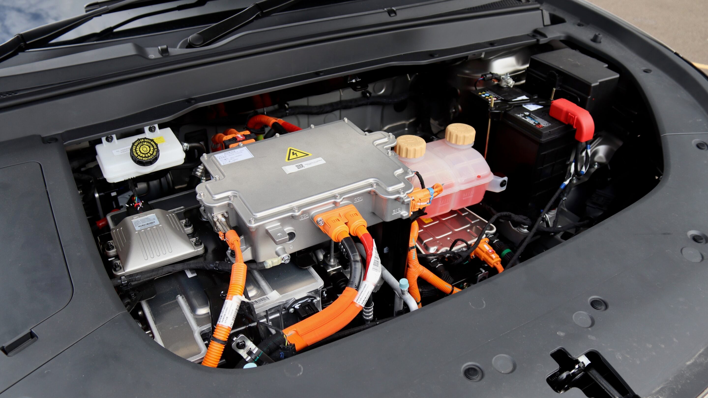Open-front view of an electric vehicle's engine bay showing a silver high-voltage power module with orange high-voltage cables and connectors.