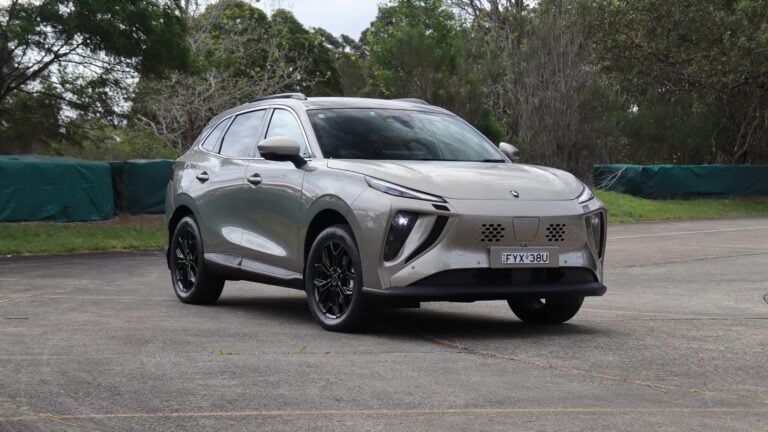Front three-quarter view of a silver crossover SUV parked on a paved lot with trees and a green barrier in the background.