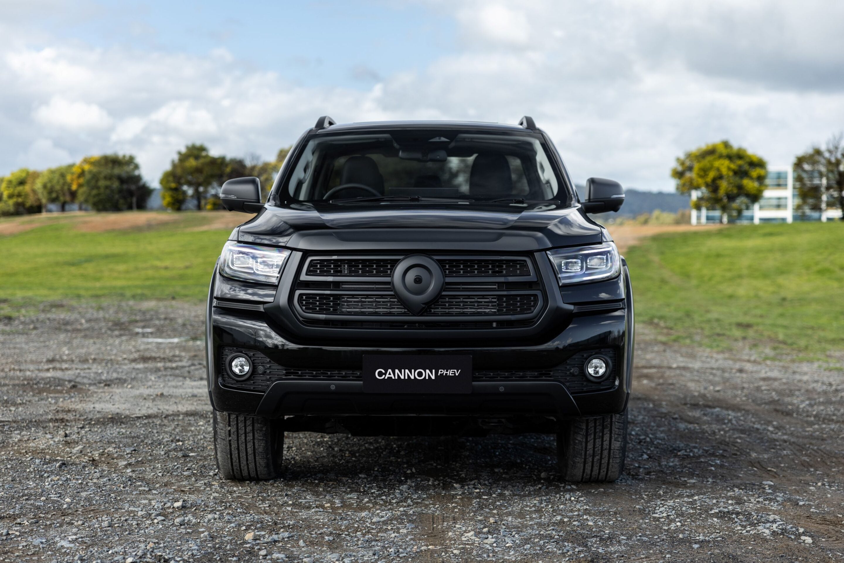 Front view of a black SUV facing the camera, parked on a gravel area with green field and trees in the background; license plate reads CANNON PHEV.