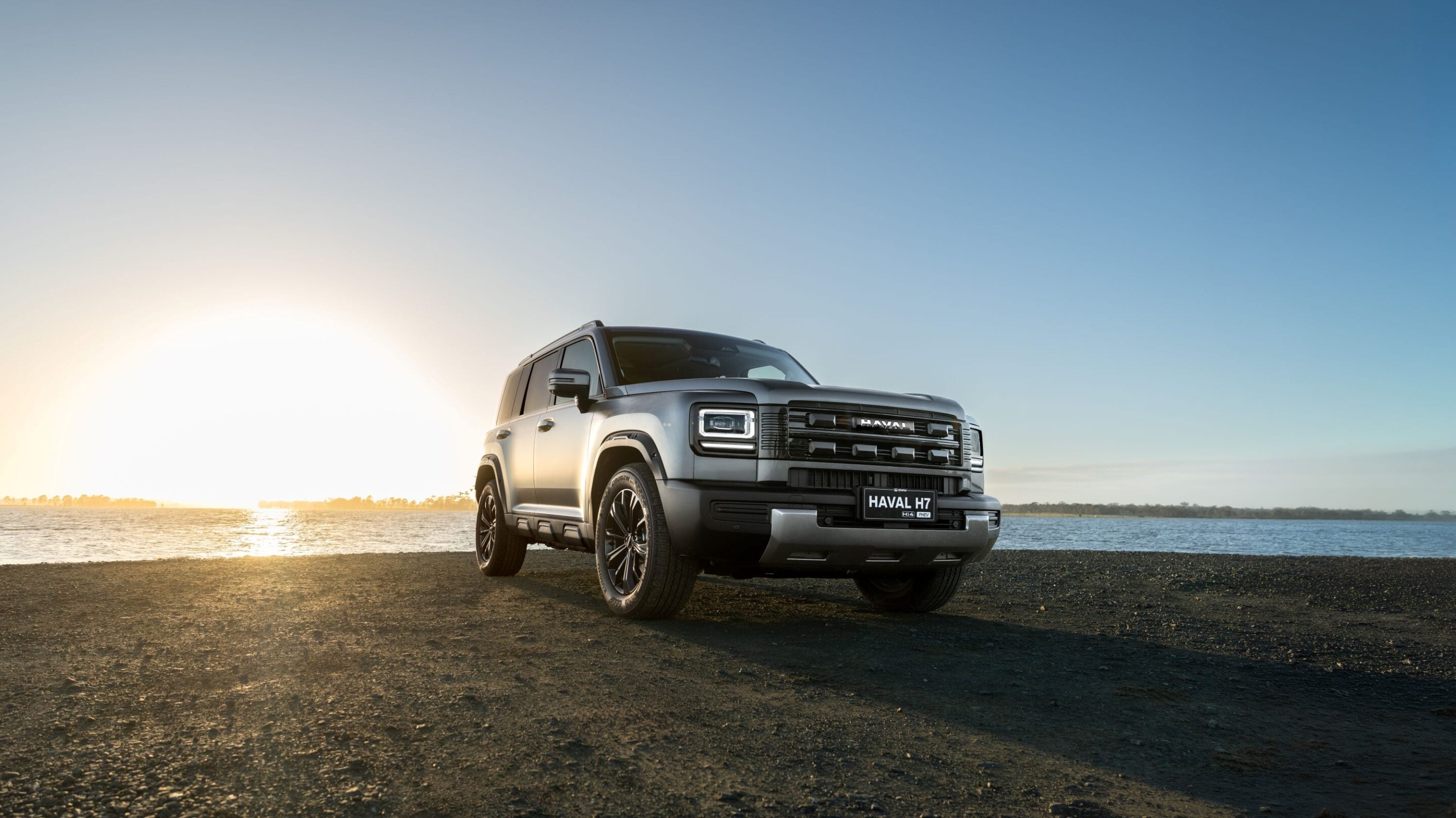 Silver Haval H7 SUV parked on a rocky shoreline at sunset with water in the background.