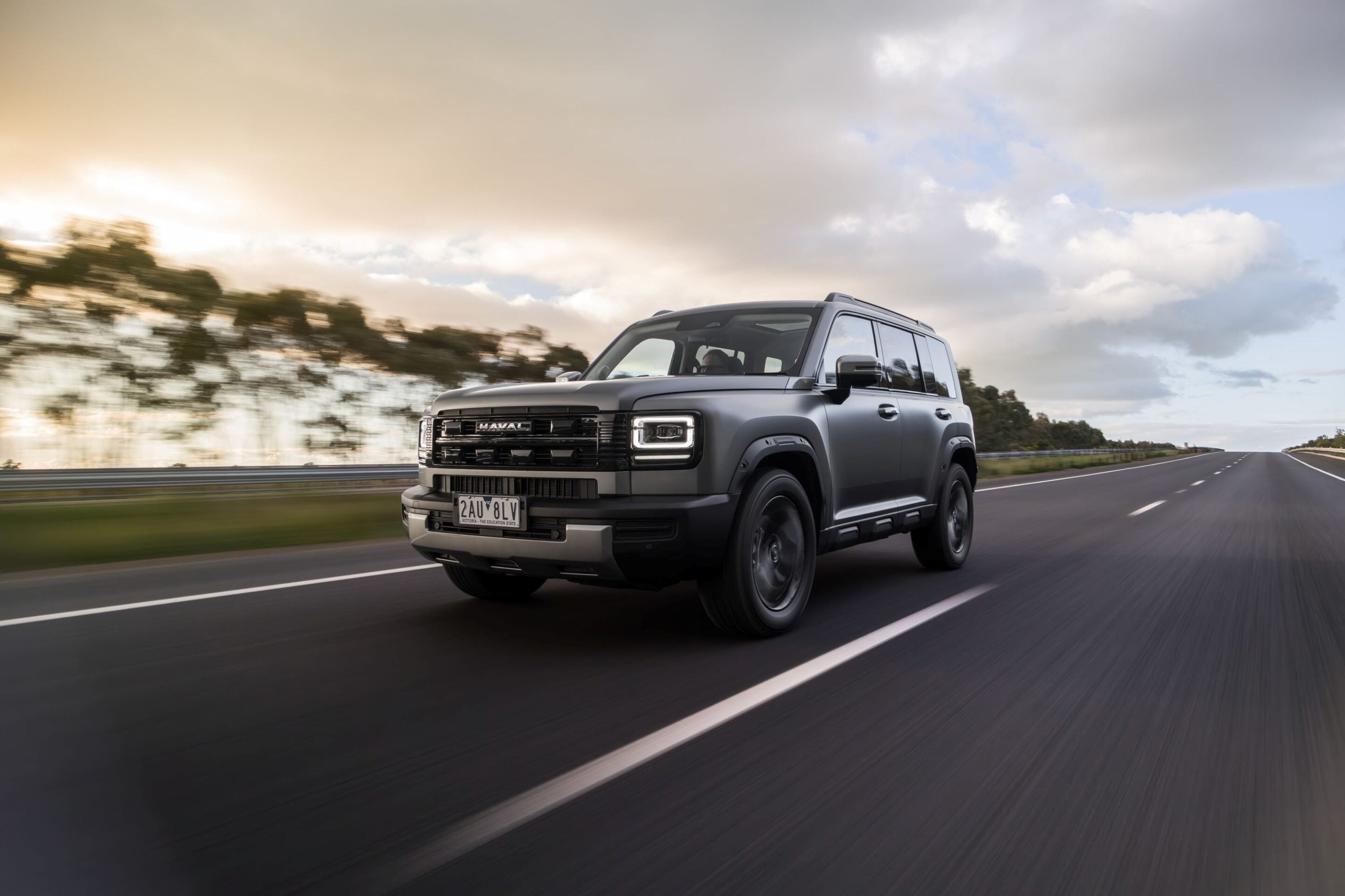 Gray SUV driving on a highway at speed with blurred trees and cloudy sky in the background, motion blur showing motion intent.