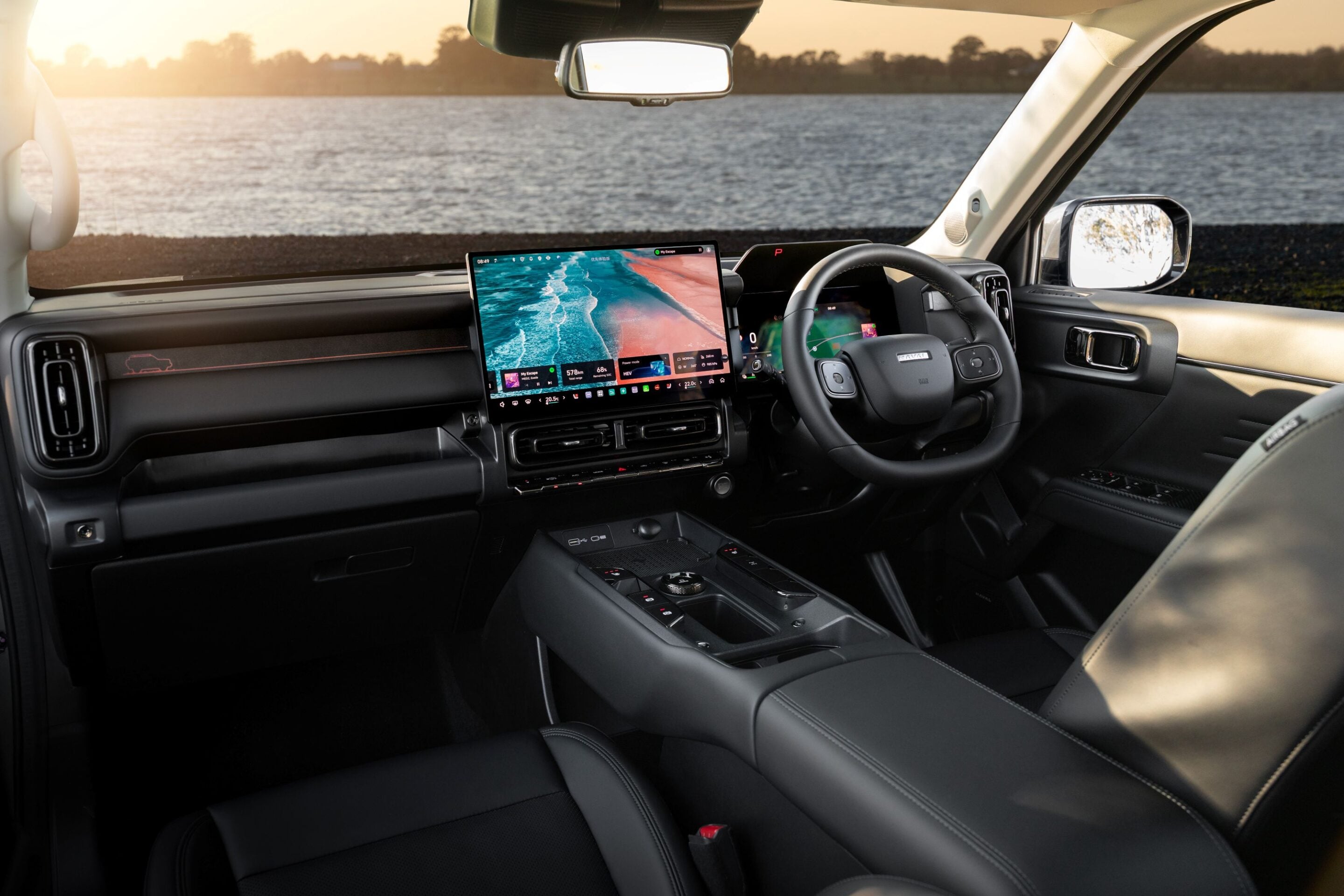 Modern car cabin with two large dashboard touchscreens, steering wheel, and sunset over a lake visible outside at the windshield edge.