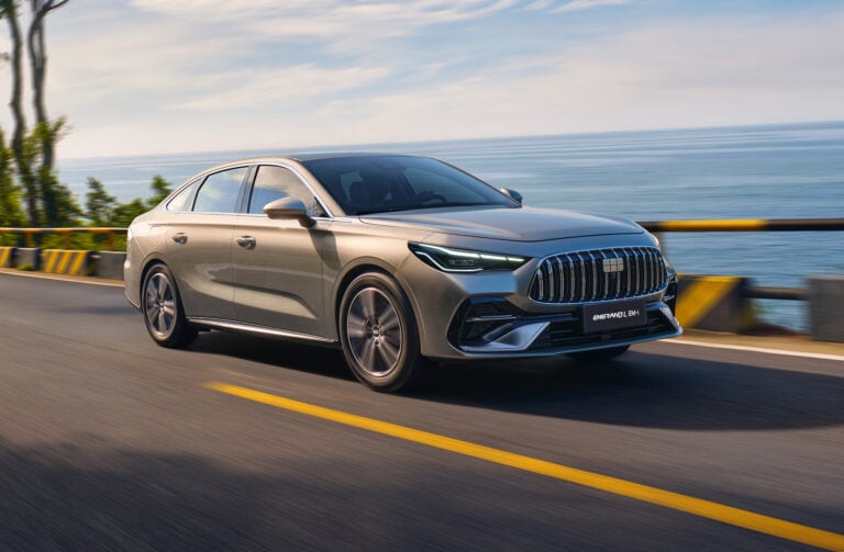 Silver sedan driving on a coastal highway with the sea in the background and guardrails on the side.