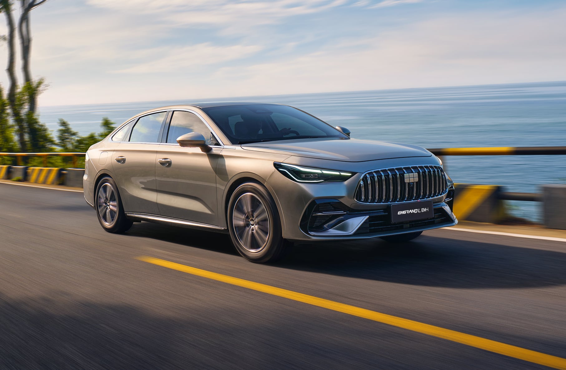 Silver sedan driving on a coastal highway with the sea in the background and guardrails on the side.