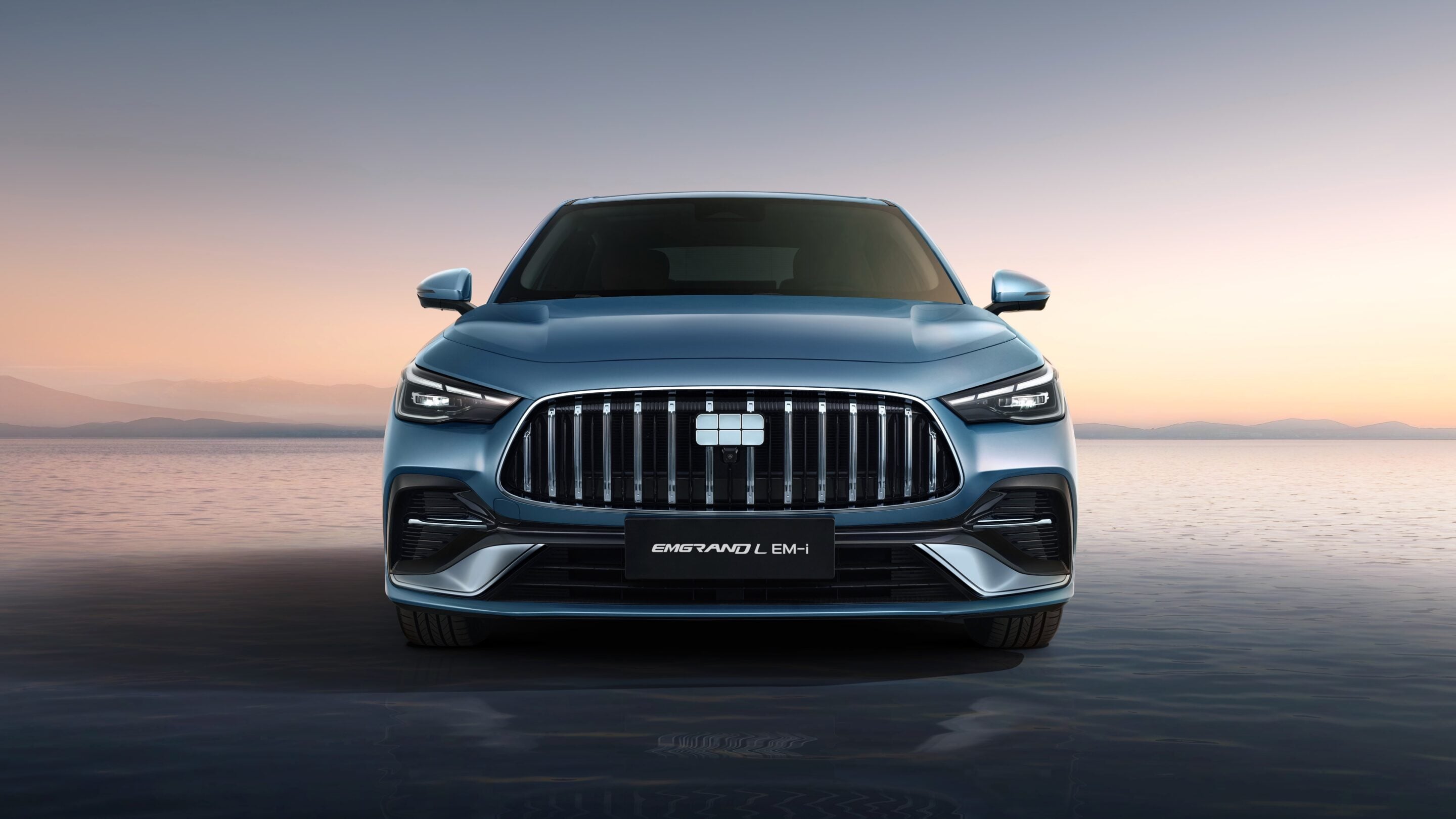 Front view of a blue EMGRAND L EM-i SUV with a prominent chrome grille at sunset by calm water.