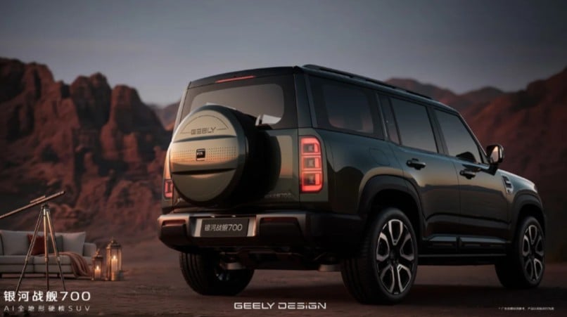 Rear view of a dark green Geely SUV with a spare-tire carrier on the back, parked in a desert canyon with a camp setup nearby.