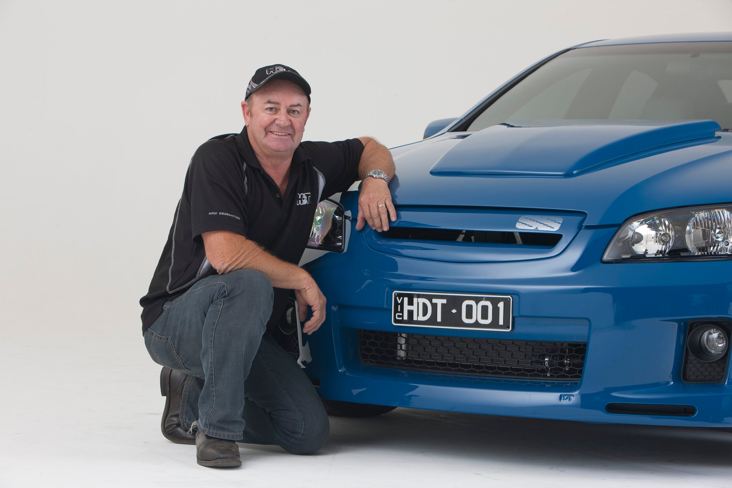 Man kneeling beside a blue car, resting his arm on the hood and smiling at the camera.