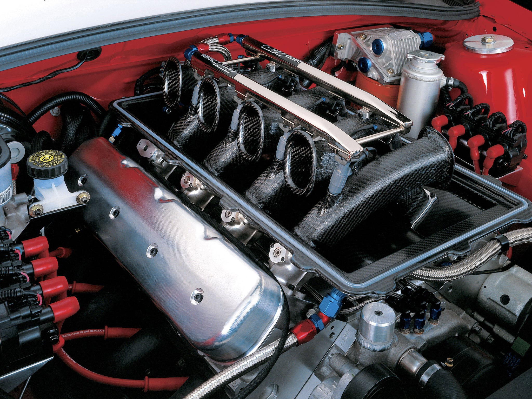 Car engine bay featuring red hoses, carbon fiber intake pipes, and polished metal strut braces with black engine components visible.