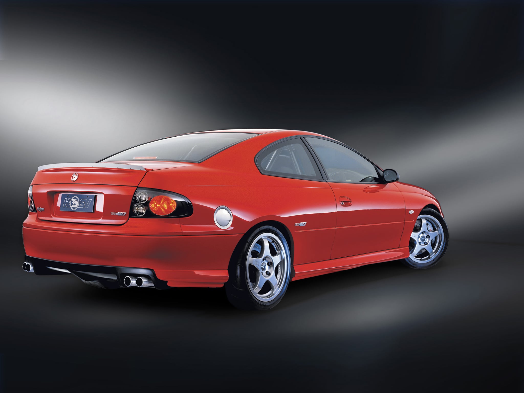Red sports car seen from the rear-right side, featuring a spoiler, chrome exhausts, and alloy wheels against a dark gradient background.