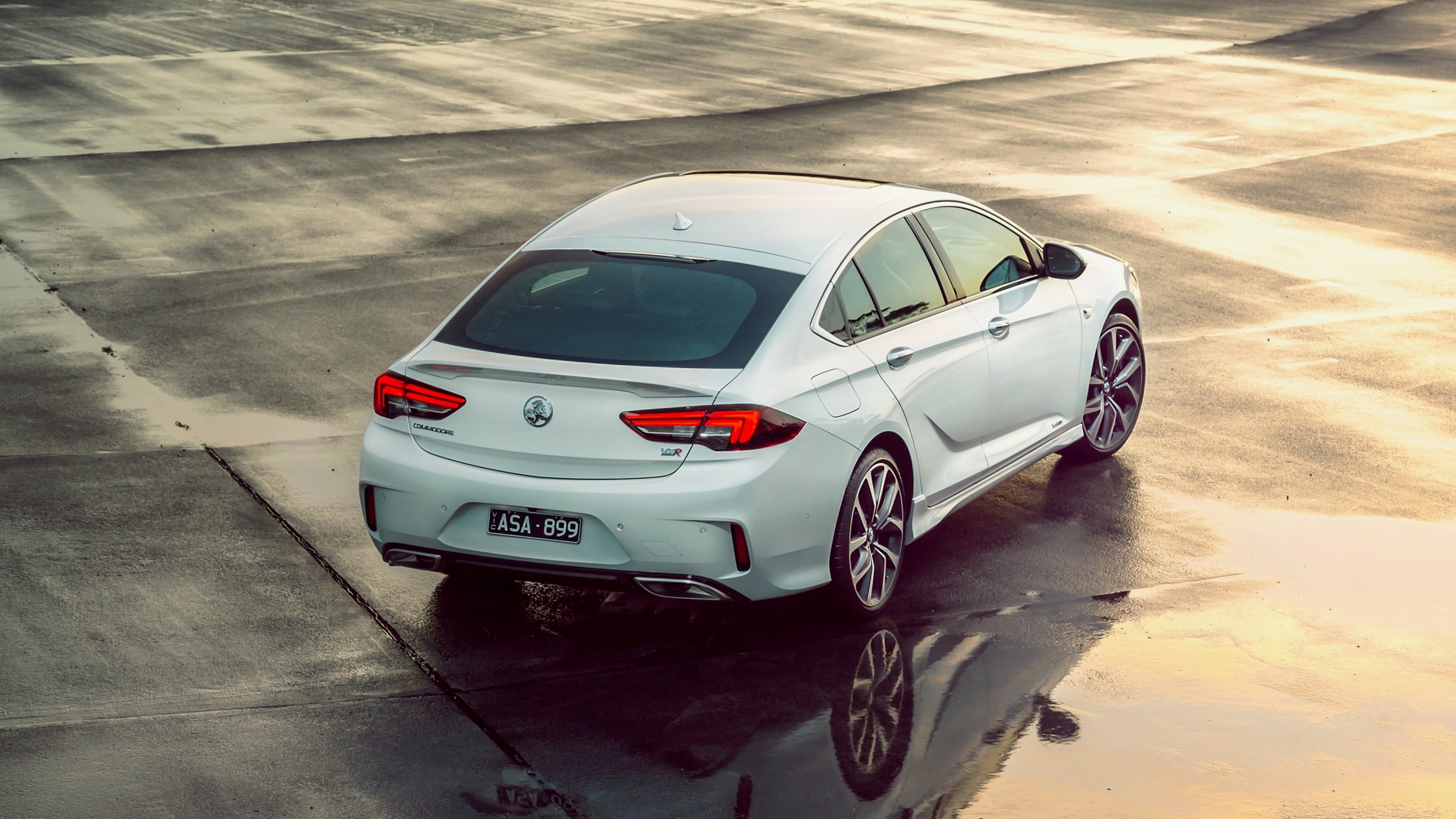 Rear three-quarter view of a white sedan parked on a wet, reflective surface at sunset