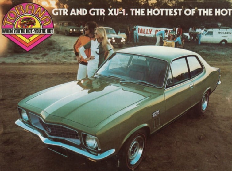 Vintage green Holden Torana GTR XU-1 on a dirt lot with two women nearby and promotional banners in the background (car show/ads).