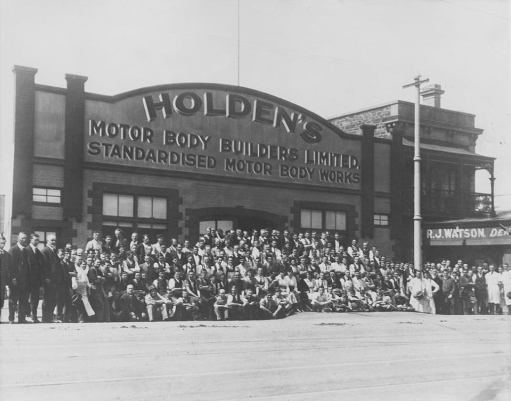 Large group of people posing in front of Holden's Motor Body Builders Limited storefront with the sign 'Standardised Motor Body Works' above the entrance.