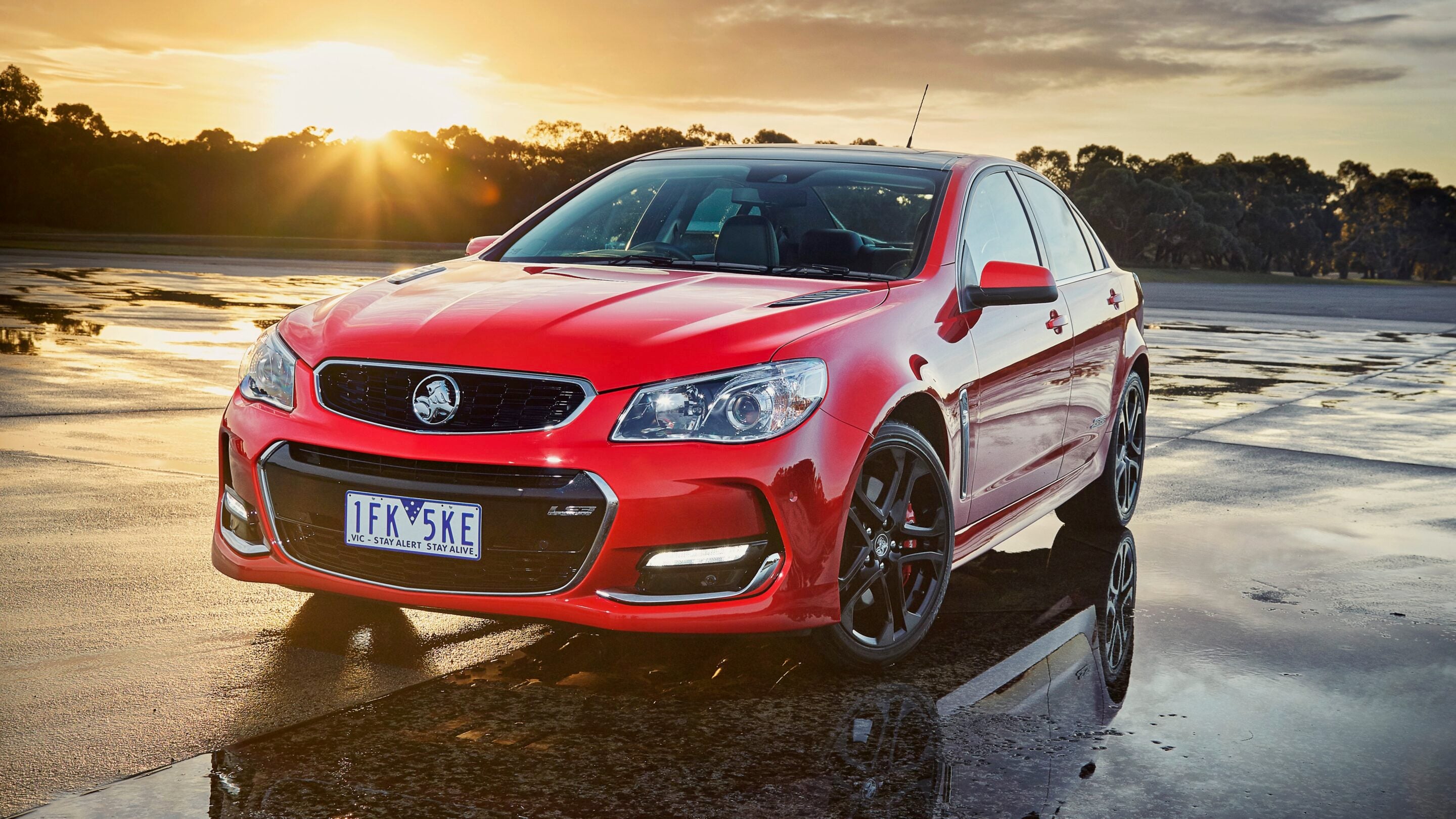 Red sedan parked on a wet surface at sunset, with reflections on the puddled ground and a tree line in the distance, license plate visible.