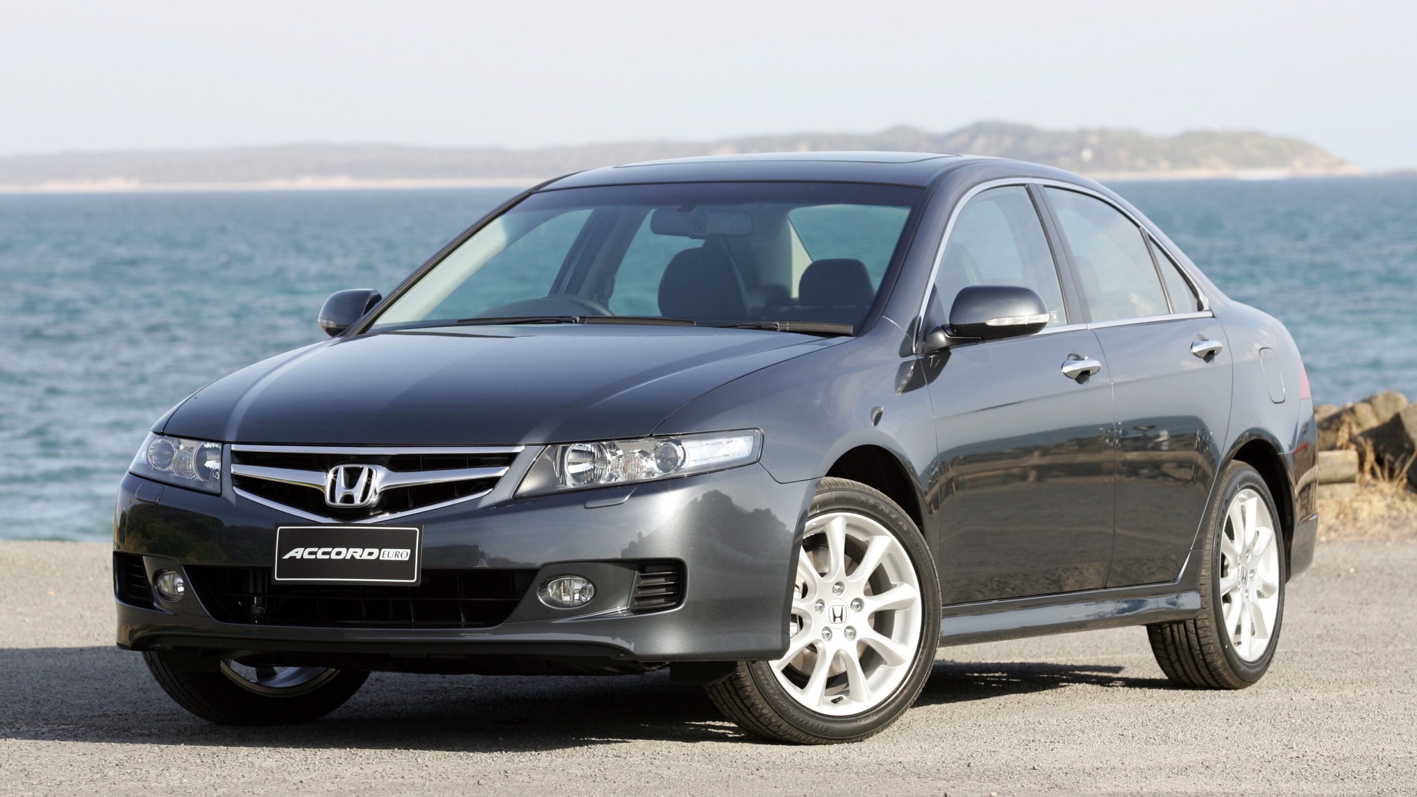 Gray Honda Accord sedan parked by a rocky shoreline with the sea in the background at a sunny day coast.
