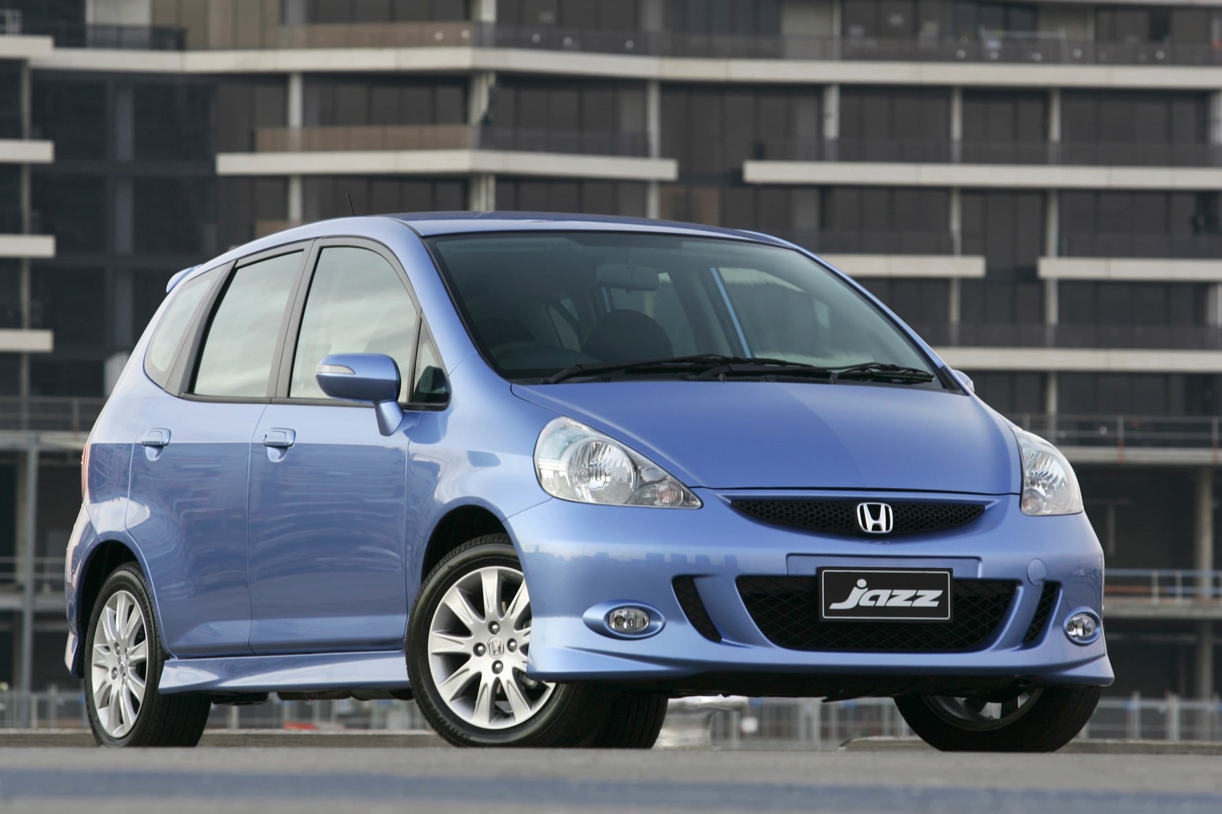 Blue Honda Jazz hatchback parked in front of a modern glass building against an urban backdrop.