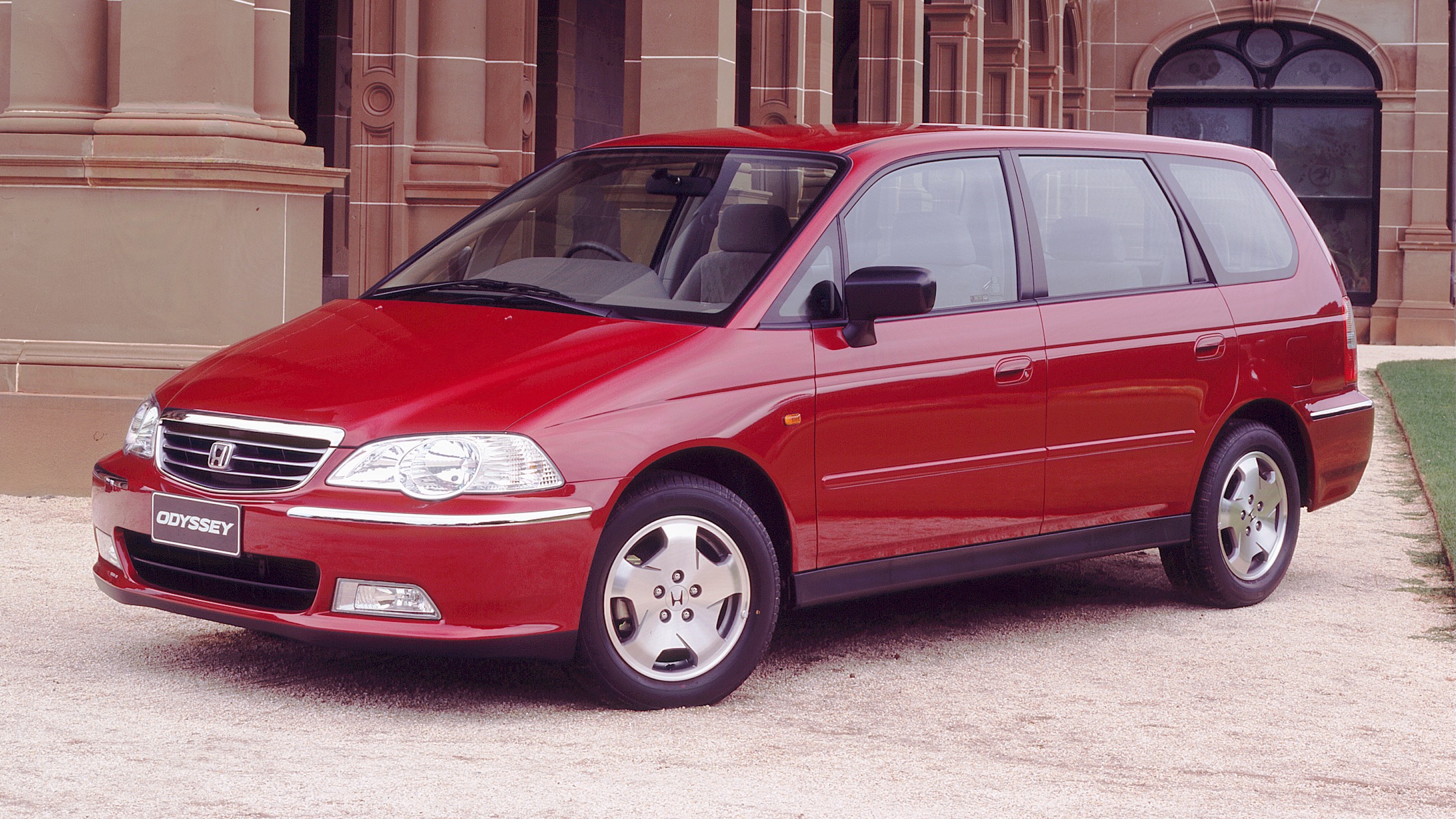 Red Honda Odyssey minivan parked on a gravel driveway in front of a beige stone building with arches and columns.