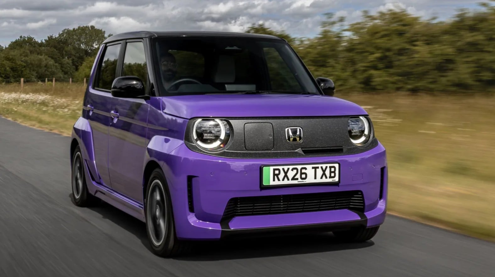 Front three-quarter view of a purple compact hatchback driving on a rural road, with round headlights and a UK license plate RX26 TXB