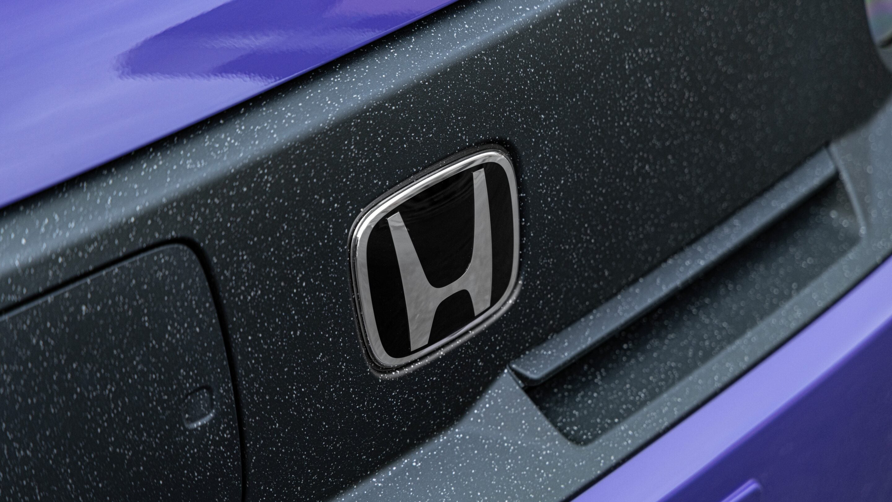 Close-up of a Honda badge on a dark car with purple trim, speckled with water droplets on the surface.