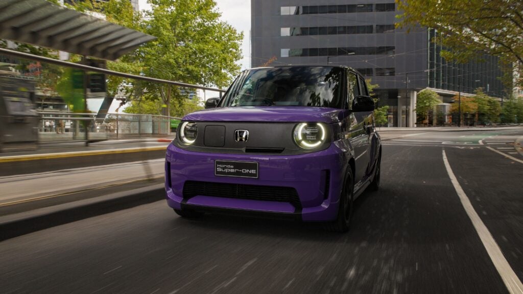 Front view of a purple Honda car driving on a city street with motion blur and glass buildings in the background, headlights on
