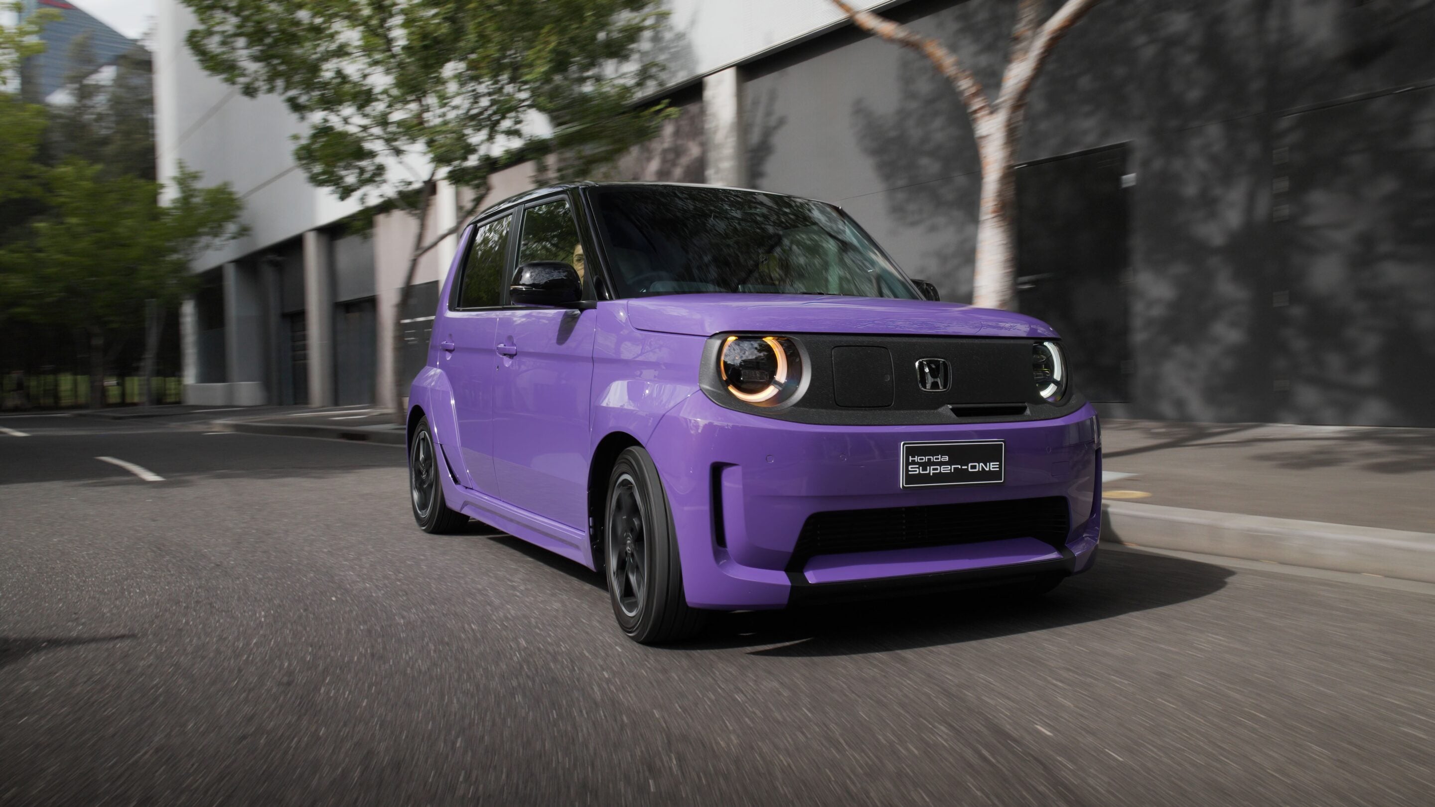 Front three-quarter view of a purple Honda car driving along a city street, with trees and a modern building in the background, headlights on as it speeds by.