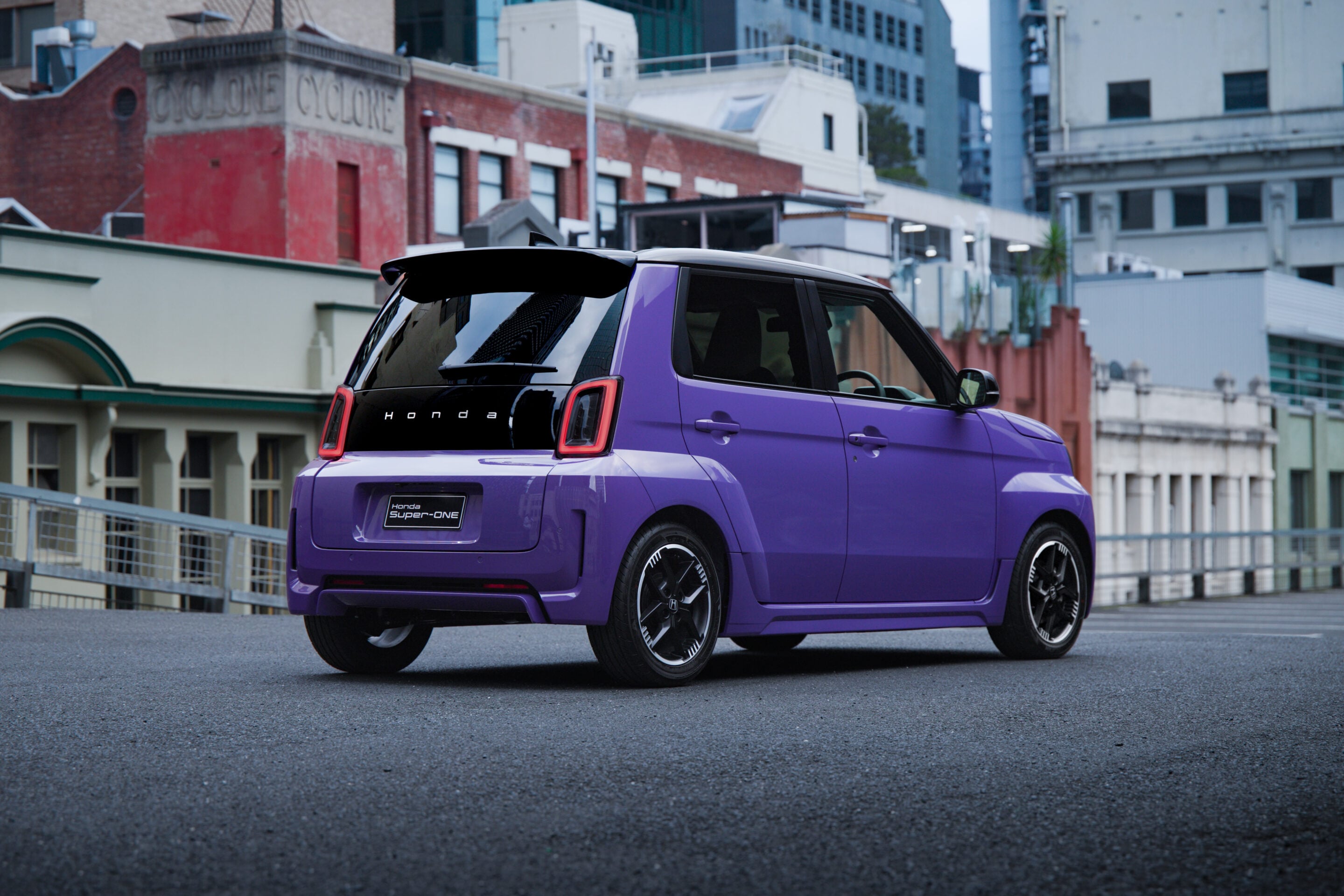 Rear three-quarter view of a bright purple Honda SUV parked on a city street with brick buildings and modern high-rises in the background, showing distinctive red taillights and dark-tinted windows.