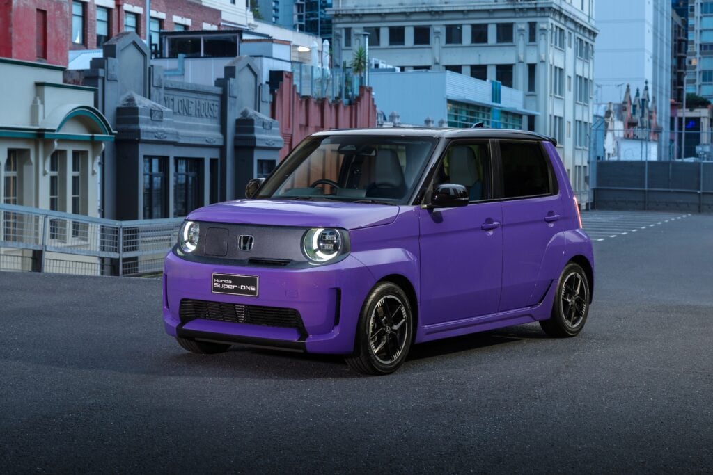 Front and side view of a purple Honda Super-ONE compact SUV parked on an urban street with colorful city buildings in the background, headlights lit.