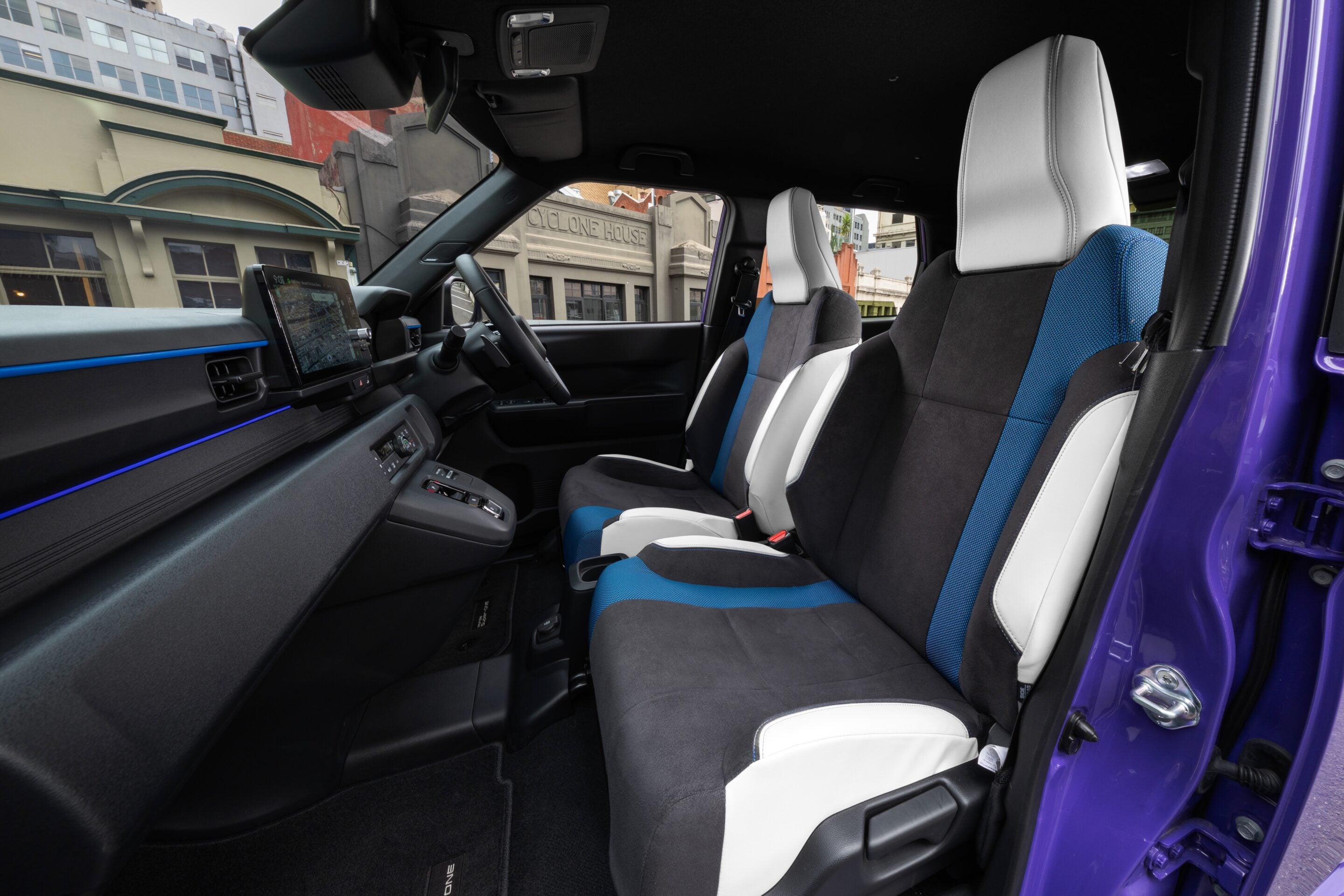 Interior of a modern car with blue-black seats and a large central touchscreen, city buildings visible through the windshield and side window in the background