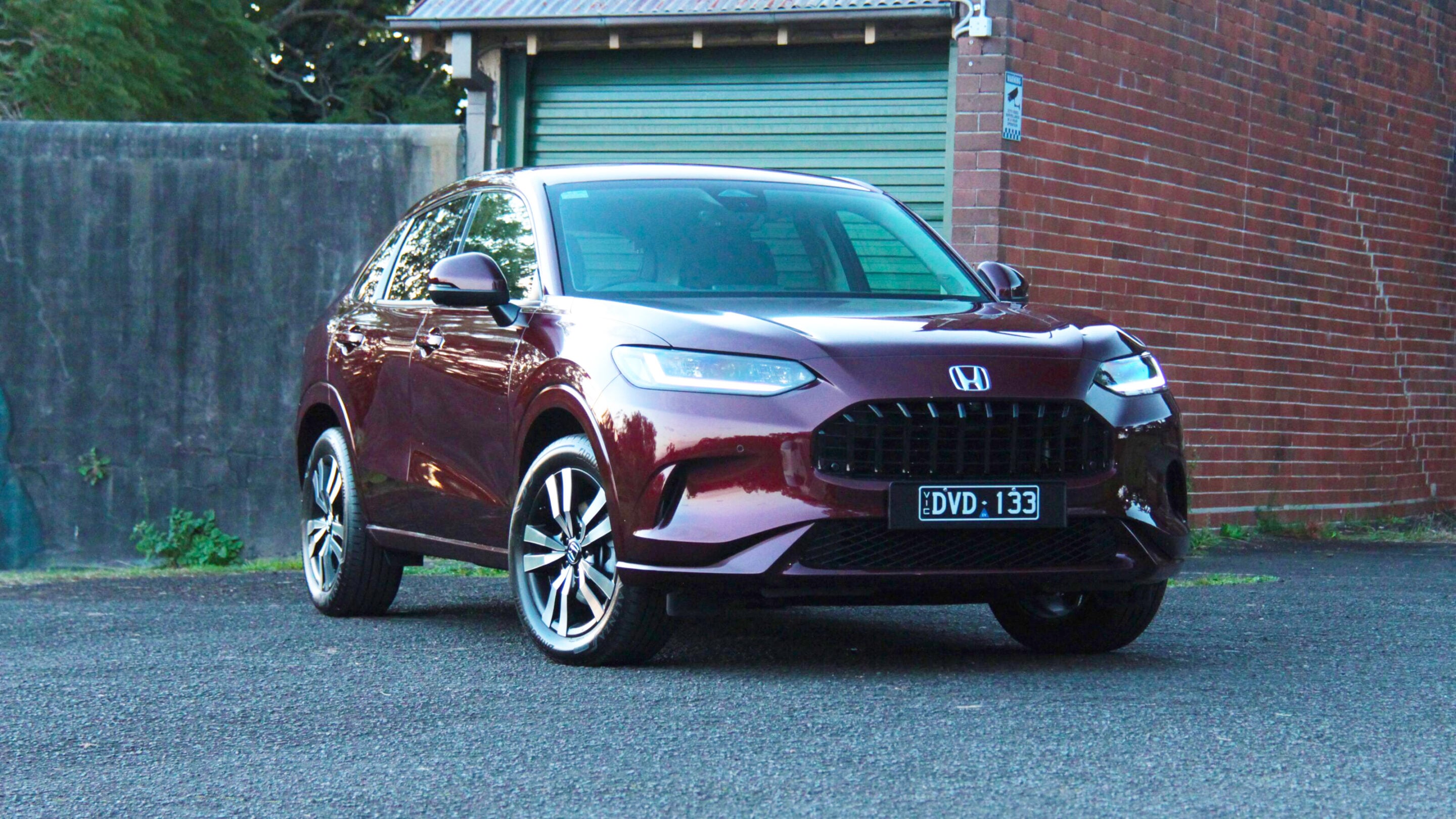 Front three-quarter view of a burgundy Honda SUV parked beside a brick wall and a green garage door behind it.