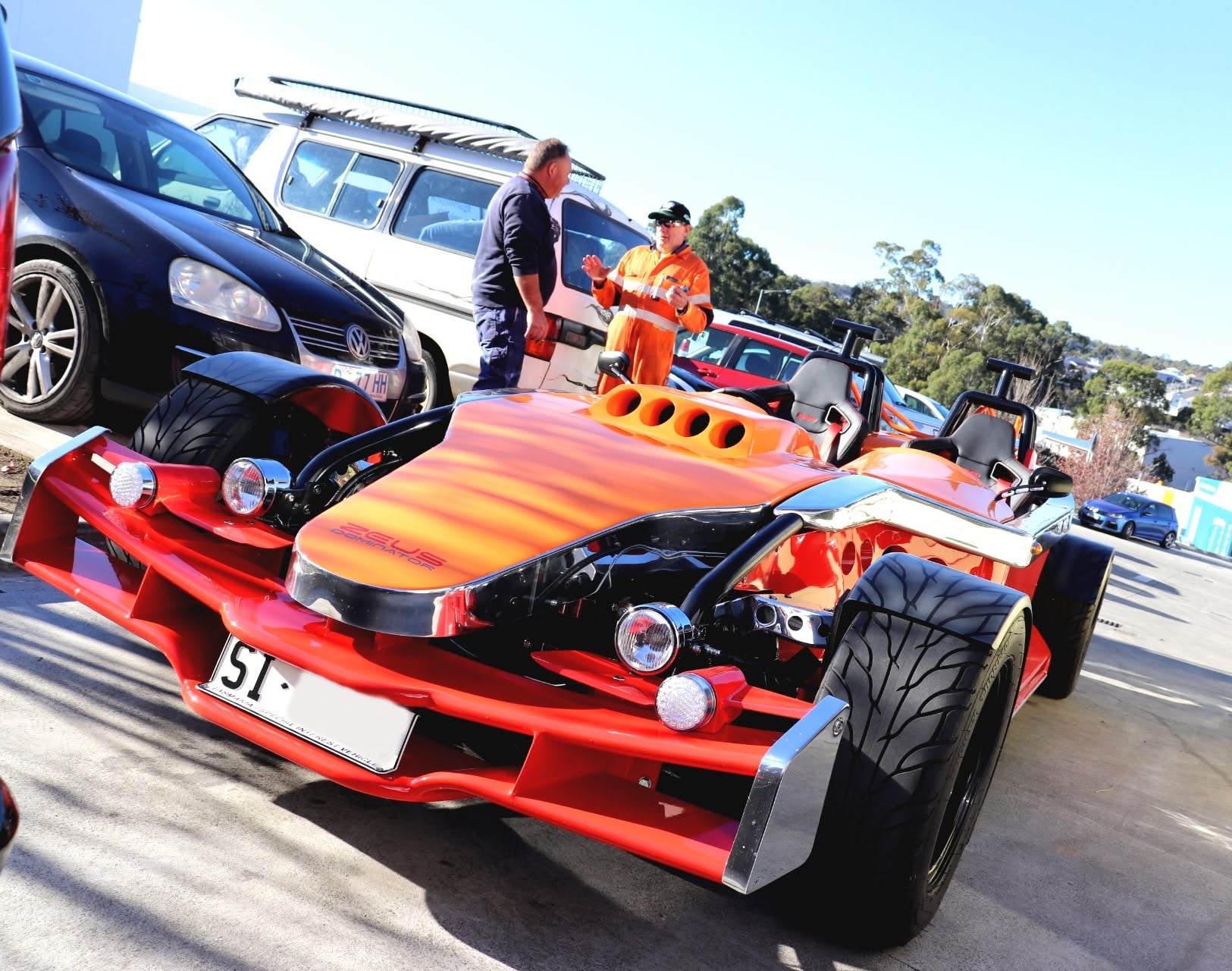 Orange open-top dune buggy with large tires in a parking lot; two men talk near a white van in the background.