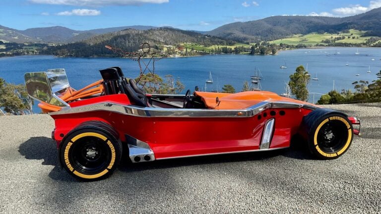 Red vintage open-top race car parked on a gravel overlook with blue lake and green hills in the background, chrome trim and large yellow-letter tires visible