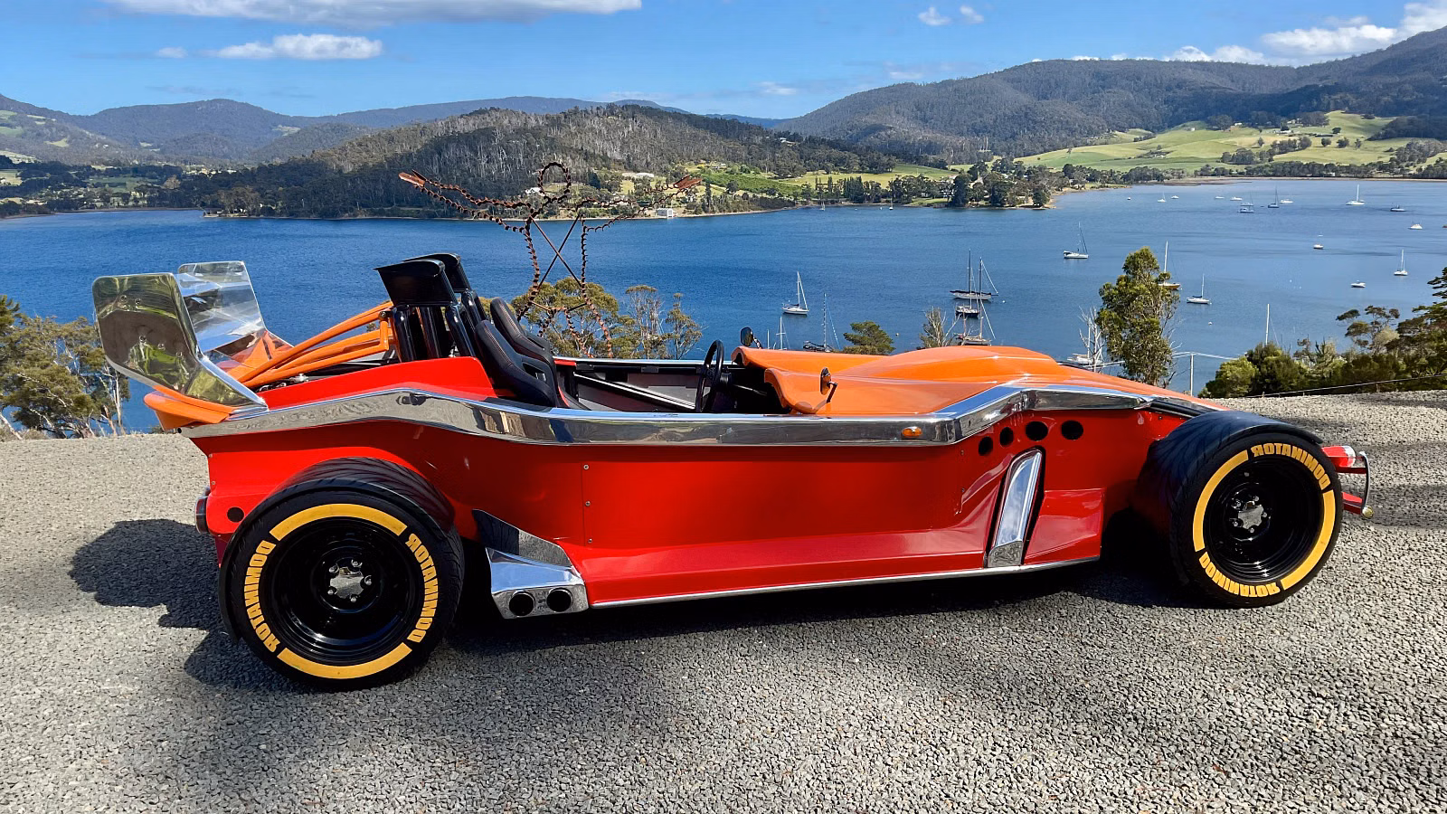Red vintage open-top race car parked on a gravel overlook with blue lake and green hills in the background, chrome trim and large yellow-letter tires visible