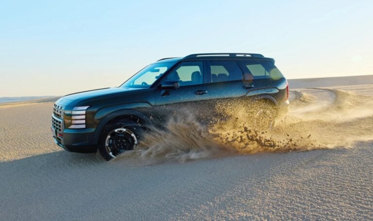 Dark blue SUV drives across a sandy desert, kicking up a spray of dust behind it at sunset-lite light.